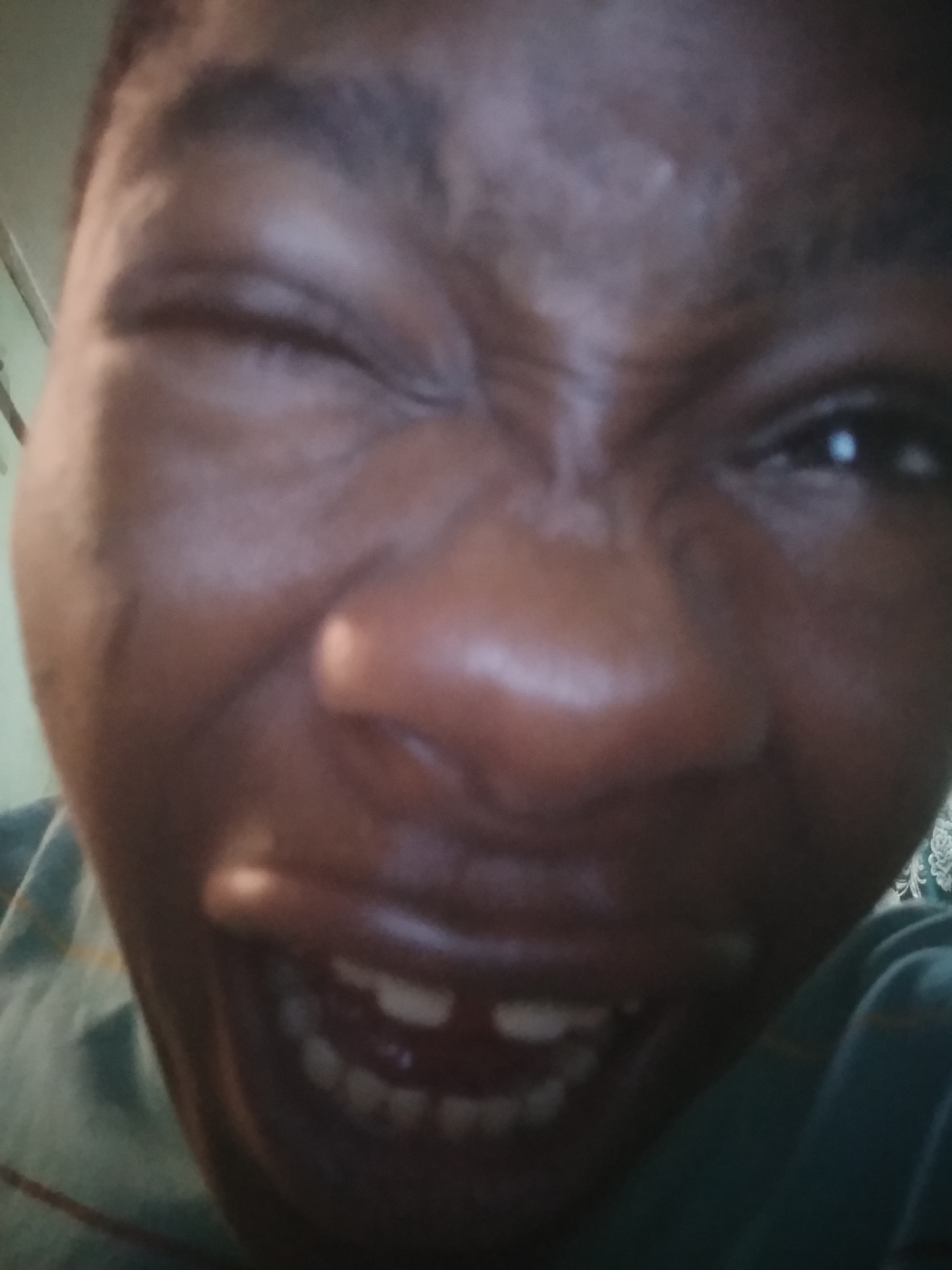 A close-up, front-facing image shows a dark-skinned person making a facial expression: One eye is open and the other is closed in a wink. The mouth is open, revealing upper and lower teeth. No specific location in Shangisha, Nigeria, is visually discernable.