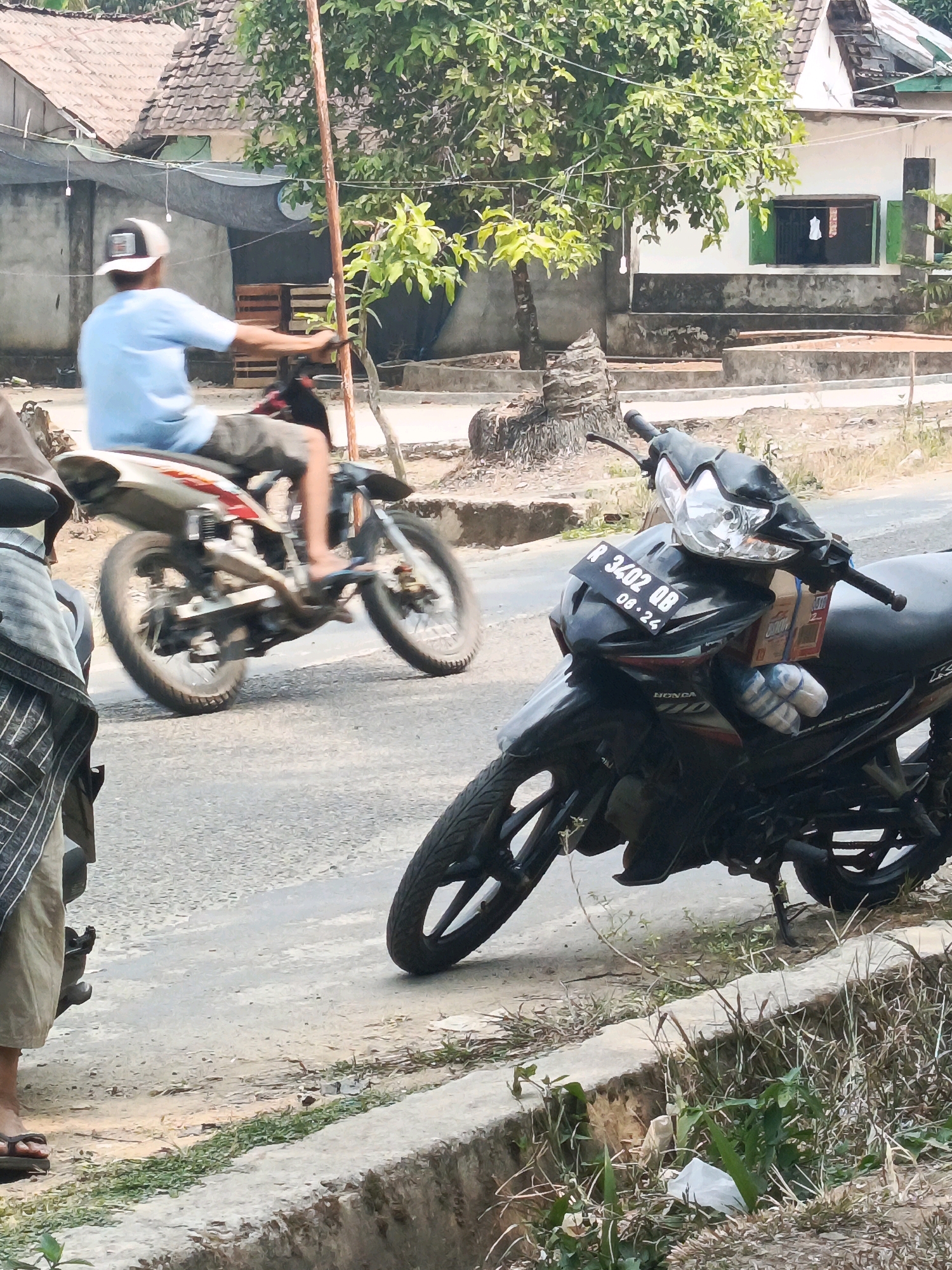 A man wearing a blue t-shirt and grey shorts rides a white and red motorcycle on a paved road. He is wearing a white and black baseball cap. The motorcycle is leaning to the left as if making a turn. In the foreground, a black motorcycle is parked on the side of the road. It has a license plate that reads "R 3402 QB 08.24". The background shows a residential area with houses and trees under a bright sky. The image appears to be taken in a street in Wonosari, Indonesia.