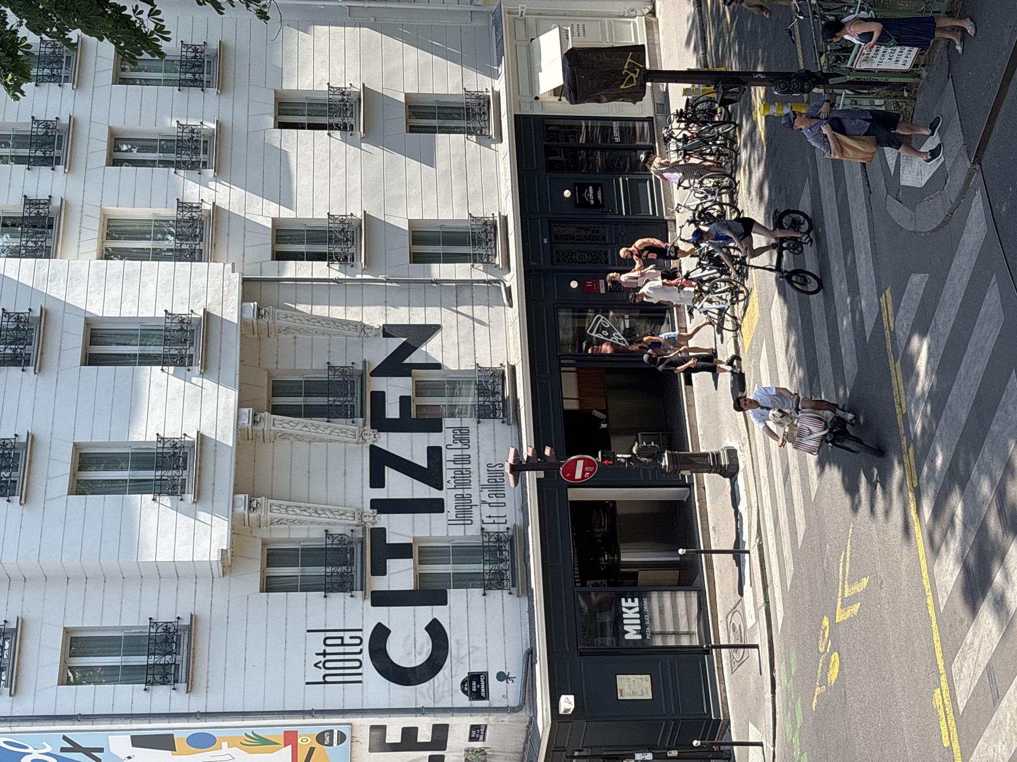 The image captures a street scene in Paris, France, likely in the Canal Saint-Martin area, given the prominent "hôtel CITIZEN Unique hôtel du Canal" signage. The focus is on the facade of the Citizen hotel and the street activity in front of it. The hotel is a multi-story white building with numerous windows featuring black wrought-iron balconies. Below the hotel, a dark-colored storefront is visible, with a sign that reads "MIKE PIZZA, SLICE, DRINKS".
On the street, pedestrians are present, with several individuals crossing a pedestrian walkway. One person is riding a bicycle carrying a dog in a basket. Another person is riding a folding bicycle. A group of people are gathered near the entrance of the MIKE establishment, and several bicycles are parked nearby. A traffic light pole is visible on the right side of the image. The overall scene depicts a sunny day with shadows cast by the buildings and foliage.