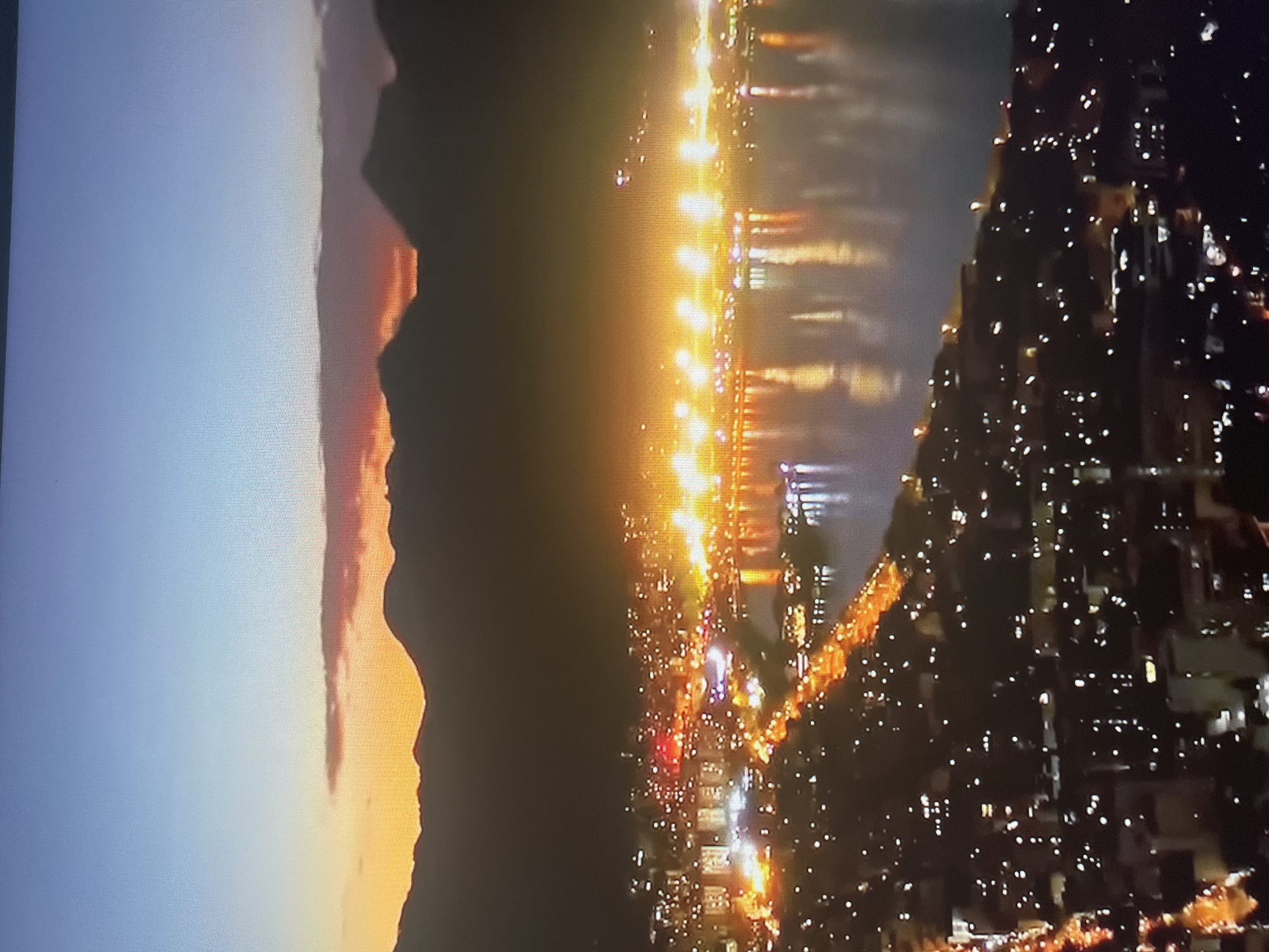 This image is an aerial view of a city at dusk, possibly captured from a high vantage point. The sky transitions from a clear, light blue at the top to a gradient of orange and red hues near the horizon, indicating the presence of a sunset. Silhouetted against the colorful sky are the dark, undulating shapes of mountains. Below the mountains, the city lights up the landscape. Numerous buildings are illuminated, creating a vibrant tapestry of white, yellow, and orange lights. A body of water, likely a bay or a lake, reflects the city's glow, creating shimmering streaks of light on its surface. Along the waterfront, a prominent line of bright lights suggests a busy commercial or recreational area. The city extends further into the foreground, with more scattered lights indicating residential areas. The overall scene conveys a sense of urban activity and beauty as nightfall approaches.