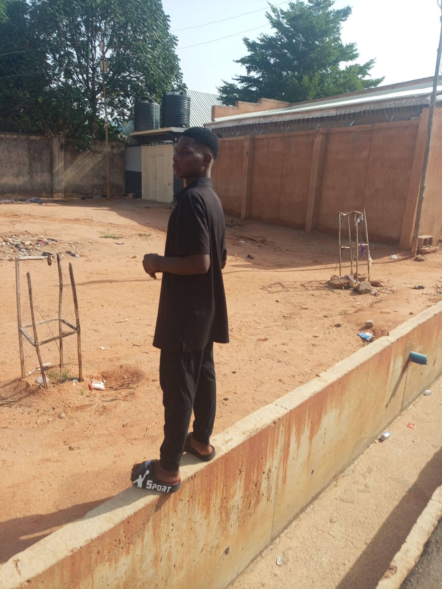 A young man stands on a low concrete wall in Tempure, Nigeria. He is wearing a dark-colored short-sleeved top, dark pants, and sandals marked with the word "SPORT". He is looking to his left. Behind him is a dirt area with scattered trash and a crude metal structure. A tall brown wall runs along the back of the area, topped with barbed wire. In the background, there are trees and water tanks. The weather appears sunny.