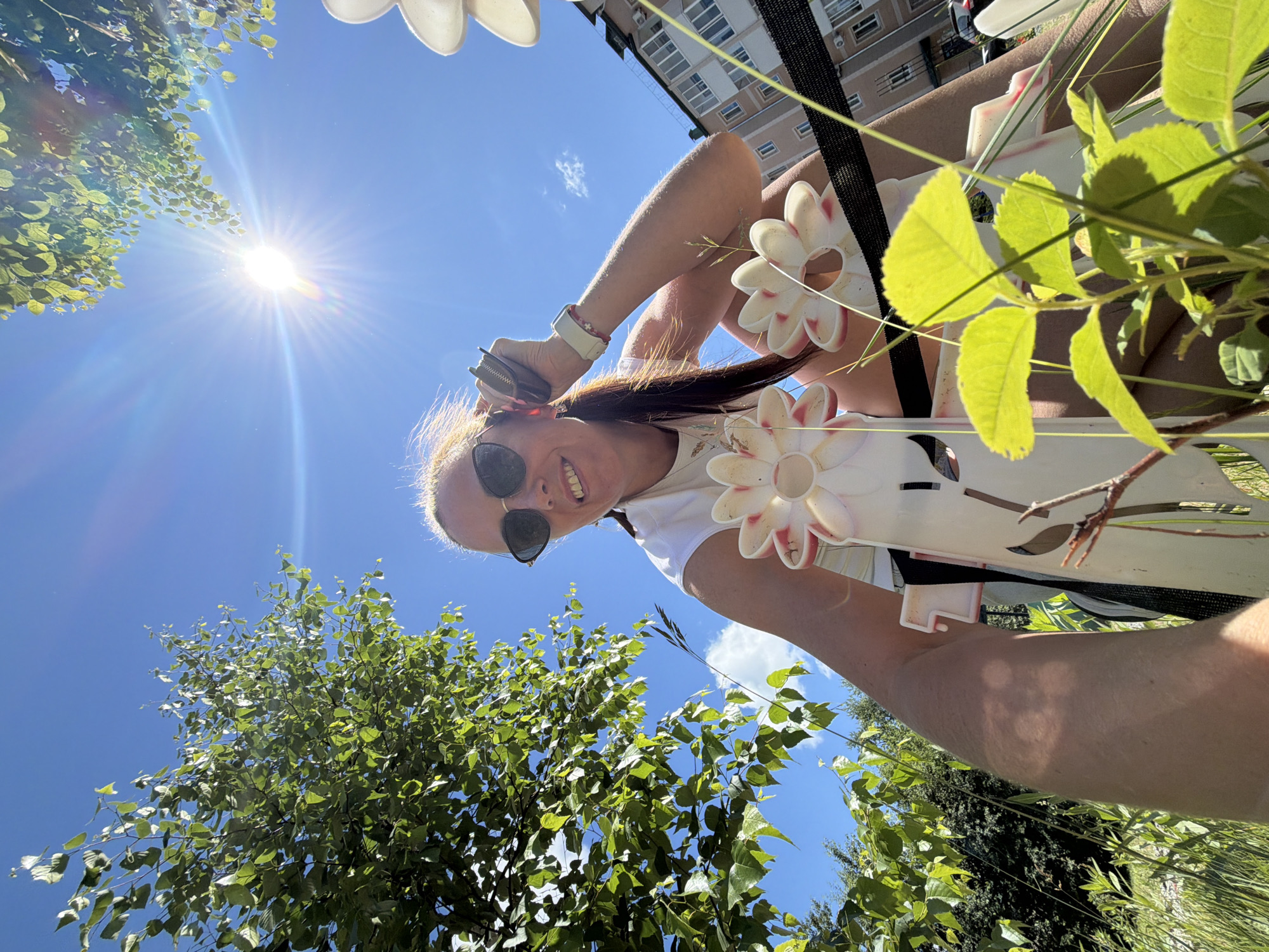 The image shows a woman in a white t-shirt and sunglasses, smiling at the camera. She has a ponytail and is wearing a watch on her left wrist. She is holding a small black wallet to her ear with her right hand. In the foreground, there are several white, flower-shaped objects with pink accents, and green foliage. In the background, there is a bright blue sky with the sun shining, and a building with windows. The overall setting appears to be outdoors, possibly in a park or garden, in a city. There is no specific indication of the location being Birobidzhan, Russia.