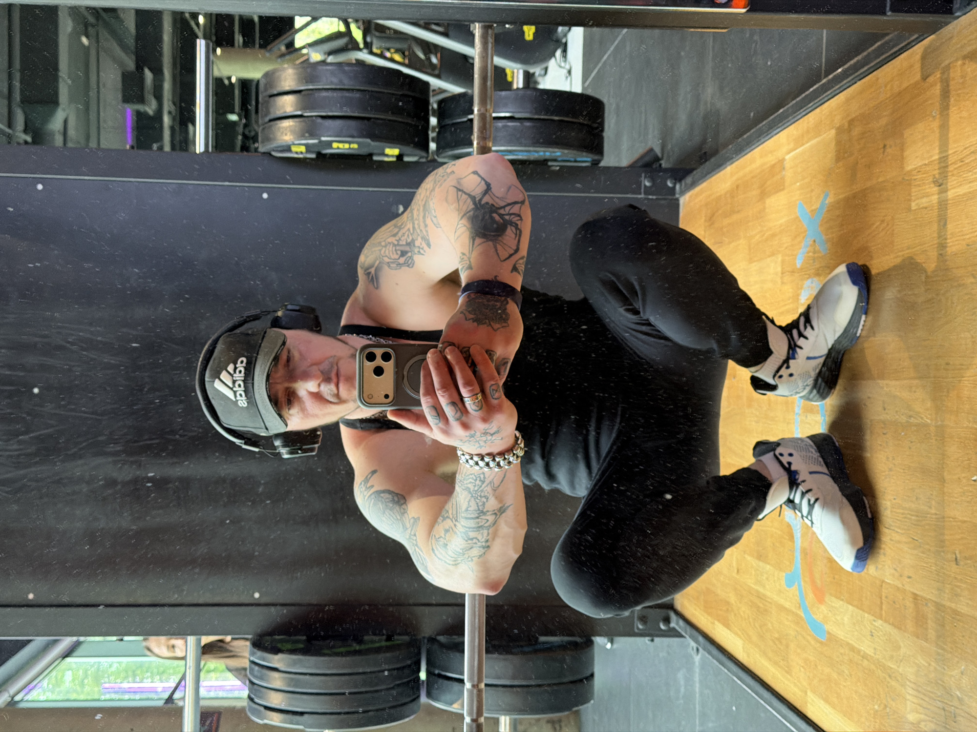 A mirrored selfie depicts a person in a gym setting. The individual, wearing a black Adidas cap and headphones, holds a smartphone, presumably to capture the image. Their attire consists of a dark tank top and black pants. Their arms are adorned with tattoos. The setting appears to be a gym with weight plates and equipment visible. The floor is a light brown wood, with blue markings. I cannot provide the location in the city of Moscow, Russia.