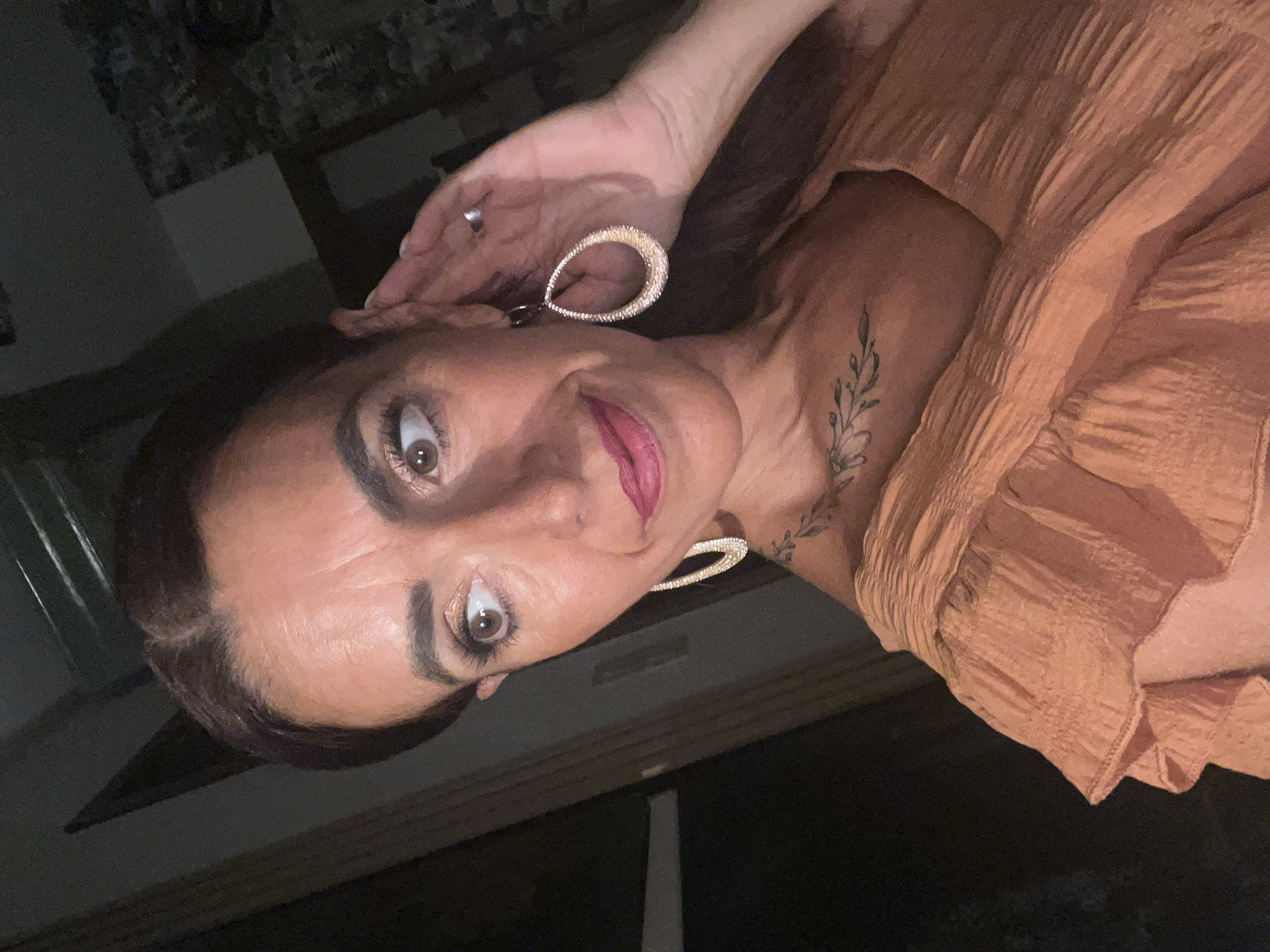 A woman, with long dark brown hair pulled back, is shown from the chest up, looking to her left with her right hand near her ear. She is wearing a peach-colored, ruffled top and large, sparkling teardrop-shaped earrings. A delicate vine tattoo is visible on her collarbone. The background is dark and indistinct, suggesting an indoor setting, possibly at night. The image appears to be a selfie. The exact location within Konz, Germany, cannot be determined from the visual information provided.