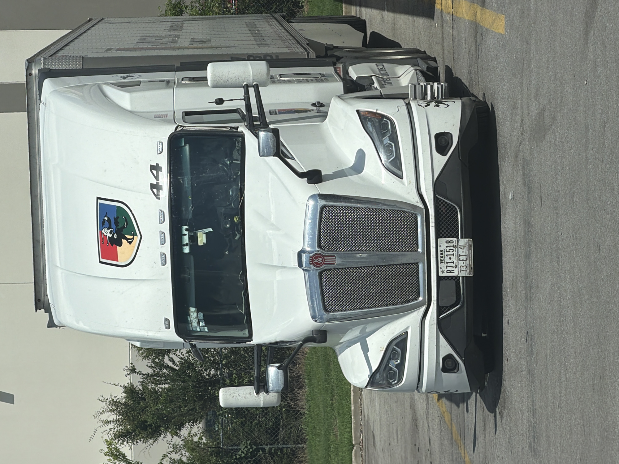A white semi-truck with a silver grille is parked on asphalt. The truck has a red Kenworth logo on the grille and a colorful shield emblem on the hood with the number 44 below it. A license plate on the front of the truck reads "TEXAS R71-1518 73-ET-4L". A large metal trailer is attached to the truck. The truck is parked in front of a light-colored building, and there are green trees and a chain-link fence behind it. The scene appears to be in an industrial or commercial area, possibly a parking lot. The location is identified as Lebanon, United States of America.