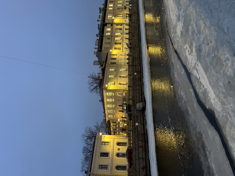 The image depicts a winter evening scene along the Moika River in Saint Petersburg, Russia. The lower half of the frame shows the canal, partially covered by white ice sheets with dark open water interspersed, reflecting warm yellow light. A stone embankment with embedded circular lights and a dark metal fence runs horizontally across the midground. A small, stone staircase descends into the water on the embankment's right side.
On the far bank, a row of multi-story, historically styled buildings with light yellow facades stands illuminated. Their windows emit warm yellow light, creating reflections on the canal's open water. Bare-branched trees are visible between and above the buildings. At least two vehicles are parked on the street behind the embankment fence: a red car on the far left and a dark-colored vehicle further right. The sky is a uniform dark blue-grey, indicating dusk or night, with a single thin, dark wire extending diagonally from the upper right towards the mid-left sky. No individuals are present in the scene.