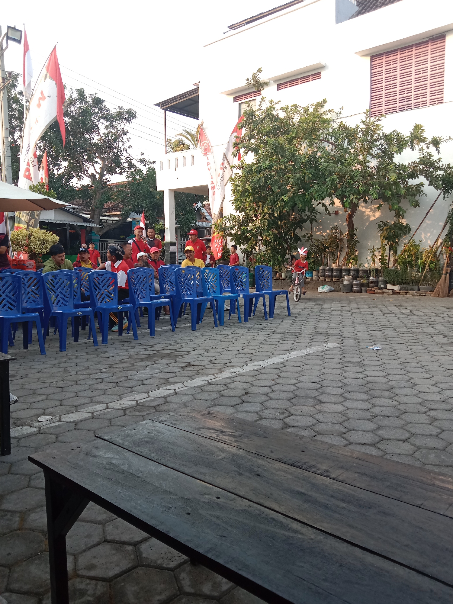 The image depicts an outdoor gathering in the city of Madiun, Indonesia, likely on a paved courtyard or lot. In the foreground, a wooden table with visible planks is positioned diagonally. The middle ground is dominated by rows of bright blue plastic chairs, some of which are occupied by people. The individuals are casually dressed, with several wearing red and white attire, suggesting a possible celebration or event. Notable are the flags displayed in the background, featuring red and white colors, and possibly Indonesian national flags or event banners. Behind the chairs and people, a white building with horizontal window slats is visible, and to the left, an awning structure shades a small area. A young person is seen riding a bicycle in the background, adding a dynamic element to the scene. The overall environment suggests a community event or social gathering taking place outdoors, possibly in the late afternoon given the lighting.