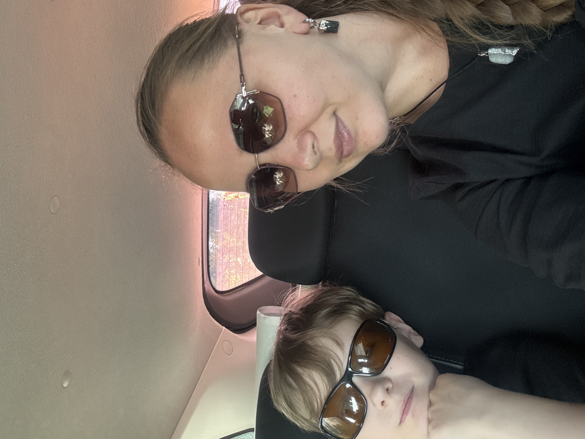 Inside a vehicle in Средний Постол, Russia, a woman and a child are visible. The woman, wearing sunglasses, a dark shirt, and a necklace, is positioned at the top of the frame. A child with sunglasses rests on the headrest below her. The interior of the vehicle is visible, including a window.
