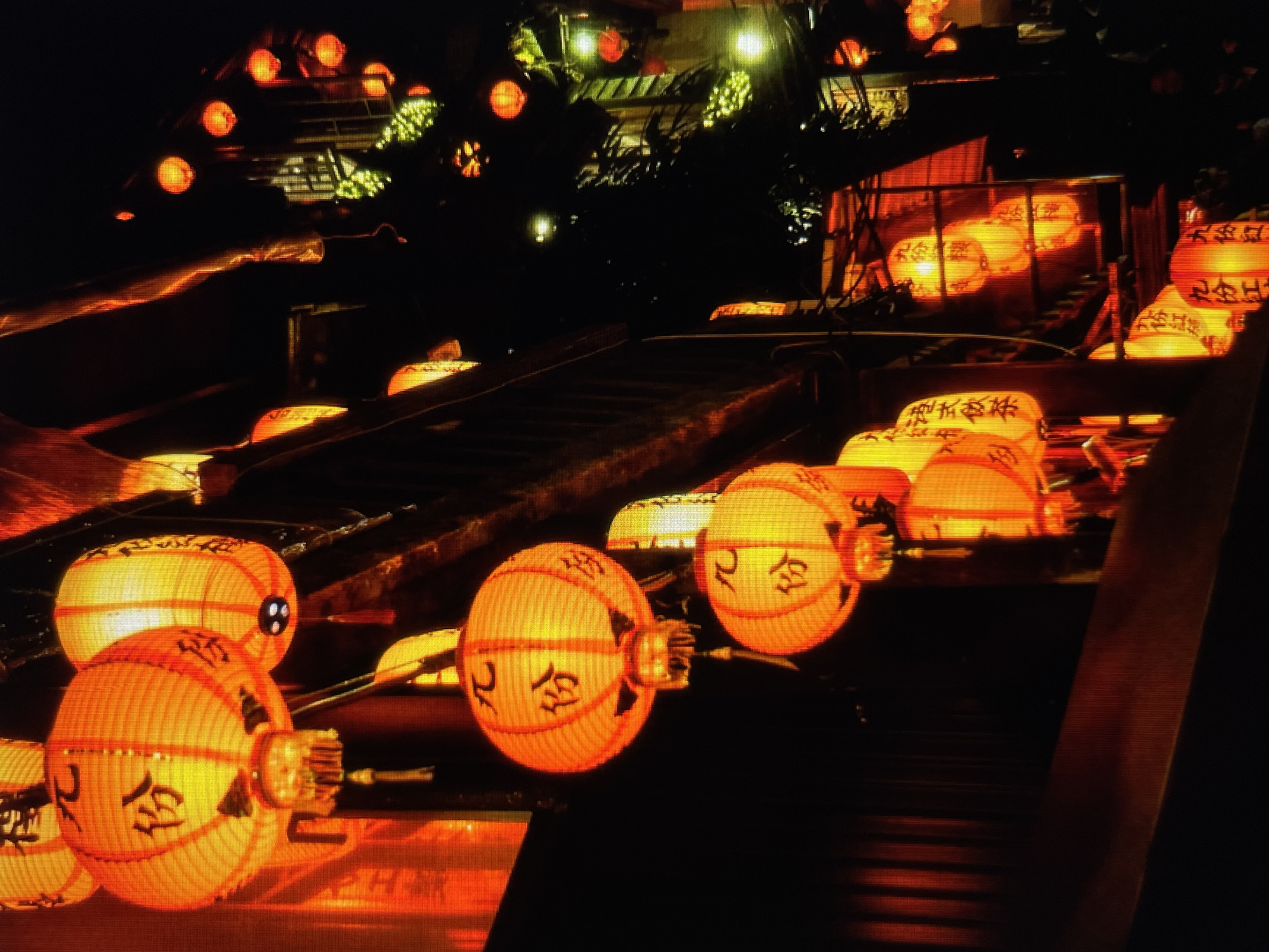 The image shows a night scene in an unidentified city, possibly in Asia, based on the presence of lanterns with Asian characters.
Subjects: The primary subjects are rows of glowing orange lanterns. Some lanterns are round and others are oval. The characters written on the lanterns appear to be Asian, potentially Chinese or Japanese.
Objects: Additional objects include structures that look like stairs, possibly part of a building or streetscape. There are also what seem to be trees or foliage visible in the upper portion of the photo. There are also a few lights at the very top of the image.
Environment: The environment is dark, suggesting a nighttime setting. The primary source of light comes from the lanterns. The scene has an urban aesthetic.