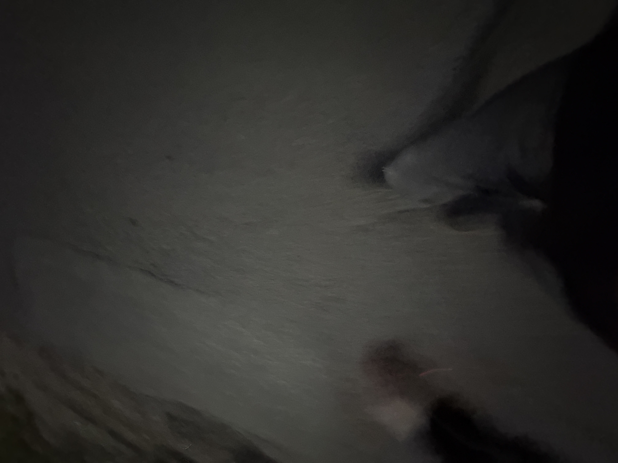 The image shows a first-person perspective of a person walking on a dark, paved surface, possibly a sidewalk or road. The lighting is dim, and the motion blur suggests movement. In the lower left of the frame, a blurred pinkish object is visible, possibly a piece of fabric or debris. To the right, a person's legs and feet are visible, clad in dark pants and a shoe. The environment is nondescript and dark, with no discernible city landmarks or features.