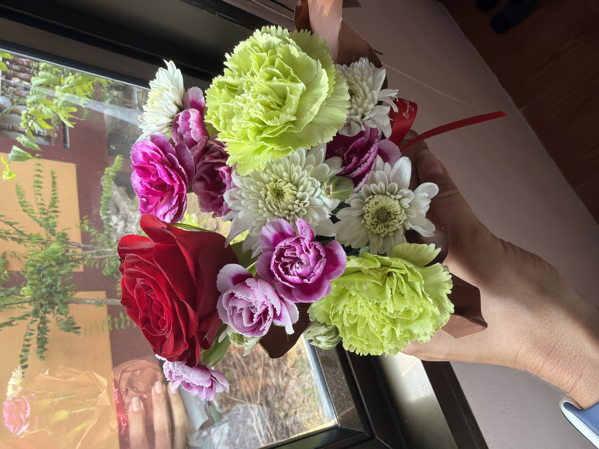 A hand holds a bouquet of flowers in front of a window. The bouquet contains a red rose, pink and purple carnations, and white chrysanthemums. The hand is tanned and has visible pores. The window overlooks trees and buildings. The scene is located in Baan Bang Rak, Thailand.