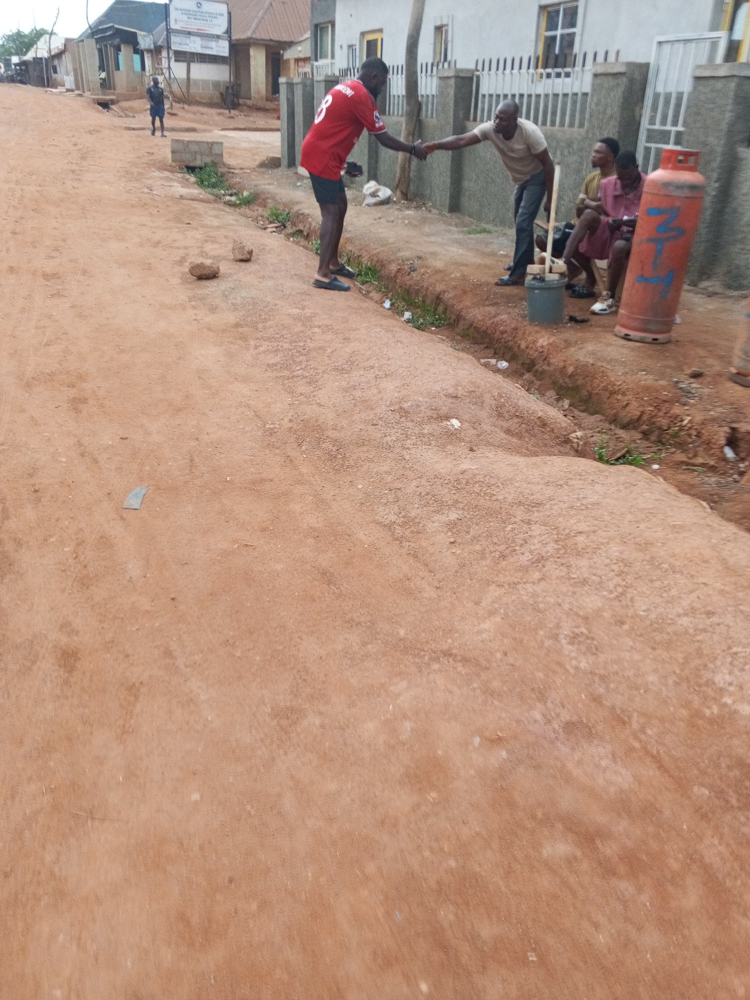 The image captures a scene on a dusty, unpaved road in a location possibly within Abuja, Nigeria. Two men are engaged in a handshake near the edge of the road. One man wears a red jersey with the number 8 and dark shorts and slippers. The other man is dressed in a light-colored shirt and dark pants, wearing sandals. Beside them, two individuals are seated on a low bench or platform. An orange gas cylinder with blue markings stands near them. A gray bucket and a wooden object are on the ground. The backdrop includes a white fence and a building. In the distance, there are other buildings along the road. The environment appears to be a residential or commercial area with basic infrastructure.