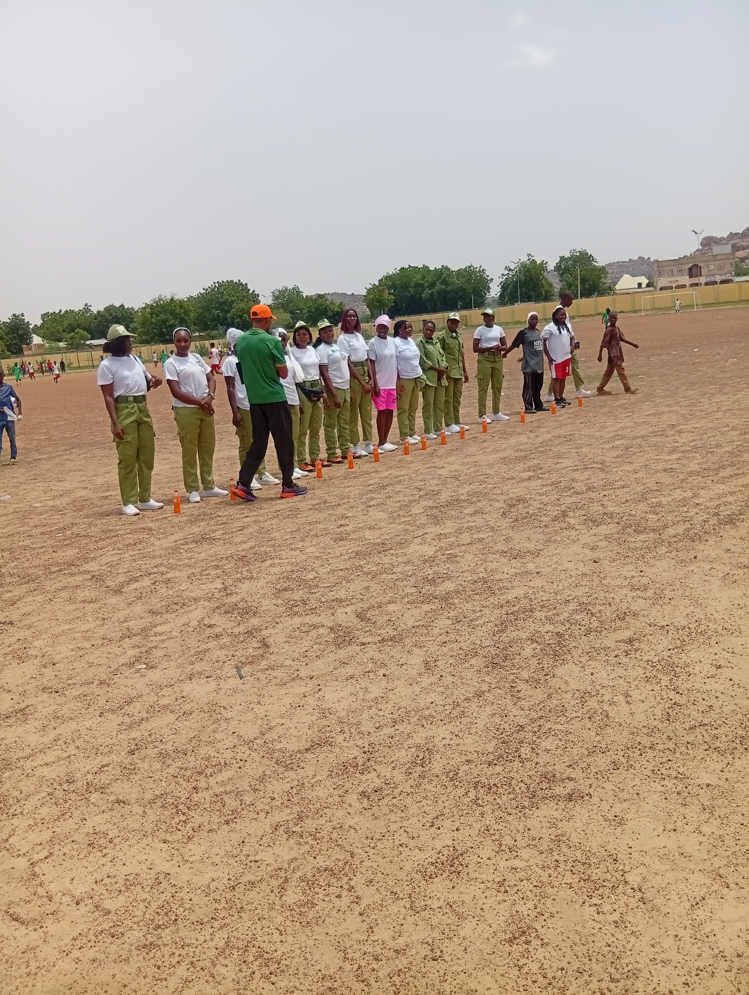 A group of people are lined up on a dusty, outdoor sports field in Dutse, Nigeria. The majority of the group appear to be young adults dressed in matching light green uniforms, consisting of shirts and pants. Some are wearing white shirts, and one individual is in a dark grey shirt and red shorts. One person in a green shirt and orange cap seems to be instructing or leading the group. The scene suggests an organized activity, possibly a training exercise or a competition, given the presence of orange cones lined up on the ground.
In the background, there are some trees, a fence, and buildings, including what looks like a stadium or sports facility with goalposts visible. The sky is overcast, indicating a cloudy or hazy day. The overall atmosphere appears to be one of mild activity and focus, with no strong indicators of overt emotion. The time of day is likely daytime, given the lighting. The ground is uneven and covered in loose soil and small pebbles, characteristic of a dusty field.