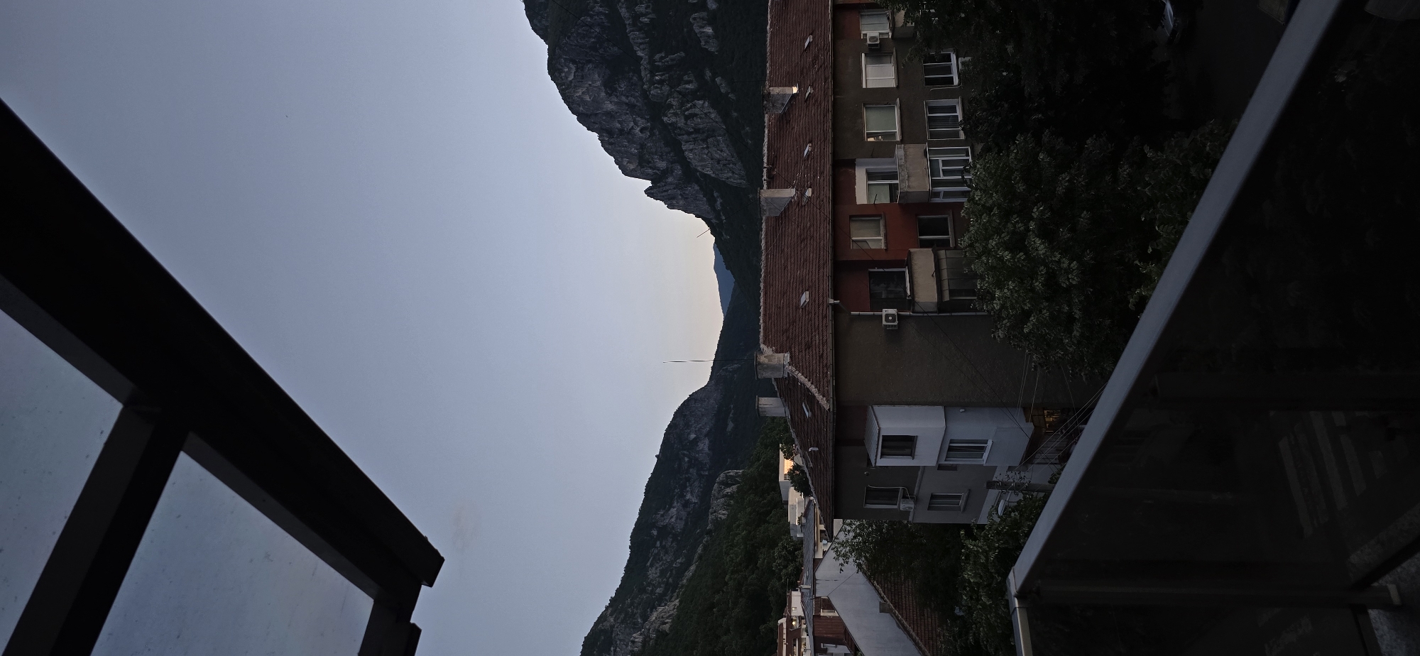 This image captures a view from a balcony overlooking a residential area and mountains at dusk or dawn in the city of Unknown, Unknown. The upper portion of the frame features a muted blue-grey sky, partially obscured by the dark, angular structure of a balcony roof or overhang in the top left. Directly below the sky, rugged, dark mountains covered in dense vegetation rise across the horizon. The mountains on the right side appear particularly steep and rocky.
In the midground, a prominent multi-story residential building occupies a significant portion of the scene. It features a reddish-brown tiled roof with multiple chimneys and a facade painted in dark grey-brown and red sections. Numerous windows, some with small balconies and exterior air conditioning units, are visible across its front. A distinct white, box-like extension protrudes from the left side of this building. To its left, other residential structures with lighter facades and reddish roofs are partially visible.
Dense, dark green foliage from trees fills the spaces between the buildings and extends up the lower slopes of the mountains. The entire lower third of the image is dominated by the dark, reflective surface of a balcony railing, likely composed of metal and glass, angled upwards from the bottom left corner. The overall lighting is dim, casting long shadows and creating a cool, subdued atmosphere. There are no people visible in the scene.