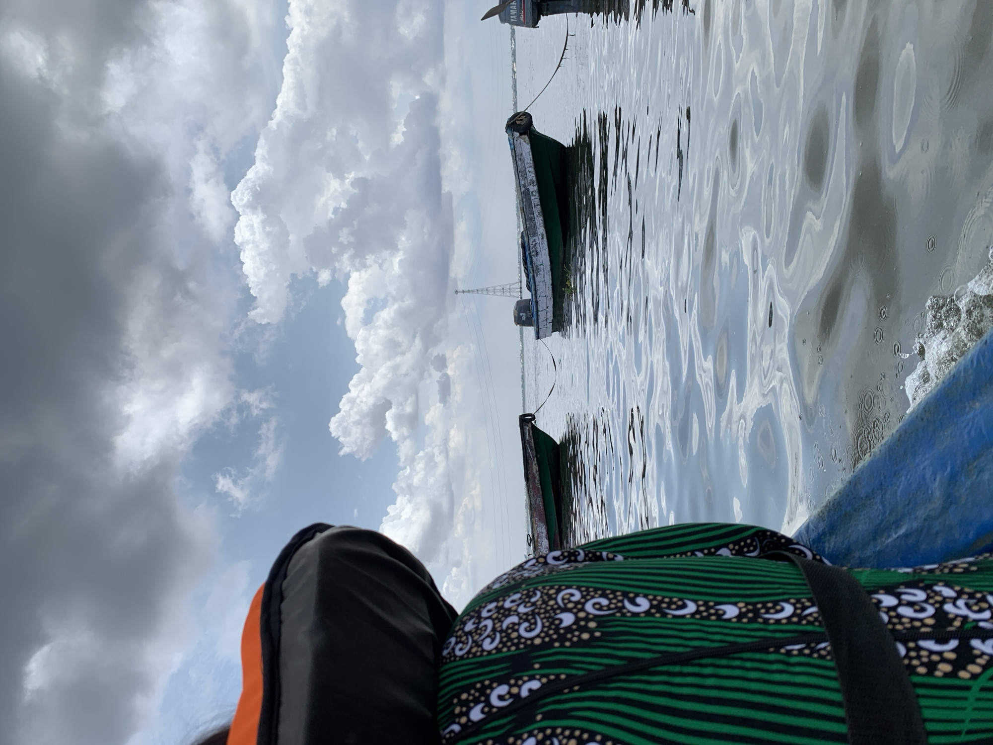 The image depicts a serene water scene from the perspective of someone seated in a boat. The original image is rotated 90 degrees counter-clockwise, showing the sky on the left and water on the right.
In the immediate foreground, the edge of a blue boat is visible on the bottom right. A person, partially seen from the chest up on the bottom left, is wearing an orange and black life vest over clothing with a distinctive green, black, and white patterned fabric, characteristic of traditional African prints.
The majority of the right side of the image is covered by a calm body of water, likely a lagoon or river in Lekki, Nigeria. The water's surface exhibits intricate ripples and reflections of the cloudy sky above. Near the foreground boat, small patches of white foam or bubbles are visible on the water.
Further into the scene, several other boats are present. A dark green boat, appearing to be a local canoe or small transport vessel, is visible in the mid-ground, partially submerged or very low in the water. In the distance, another boat features an outboard motor with the brand name "YAMAHA" clearly legible. Beyond the boats, a tall metal structure, possibly an electricity pylon or communication tower, stands against the horizon.
The sky, dominating the left portion of the image, is a significant feature. It presents a dynamic mix of large, fluffy white cumulus clouds against lighter blue patches, contrasted with darker, heavier grey clouds towards the far left. This suggests an overcast or partly cloudy daytime, with potentially varying light conditions.
The overall atmosphere is tranquil, capturing a moment of travel or repose on the water under a dramatic sky.