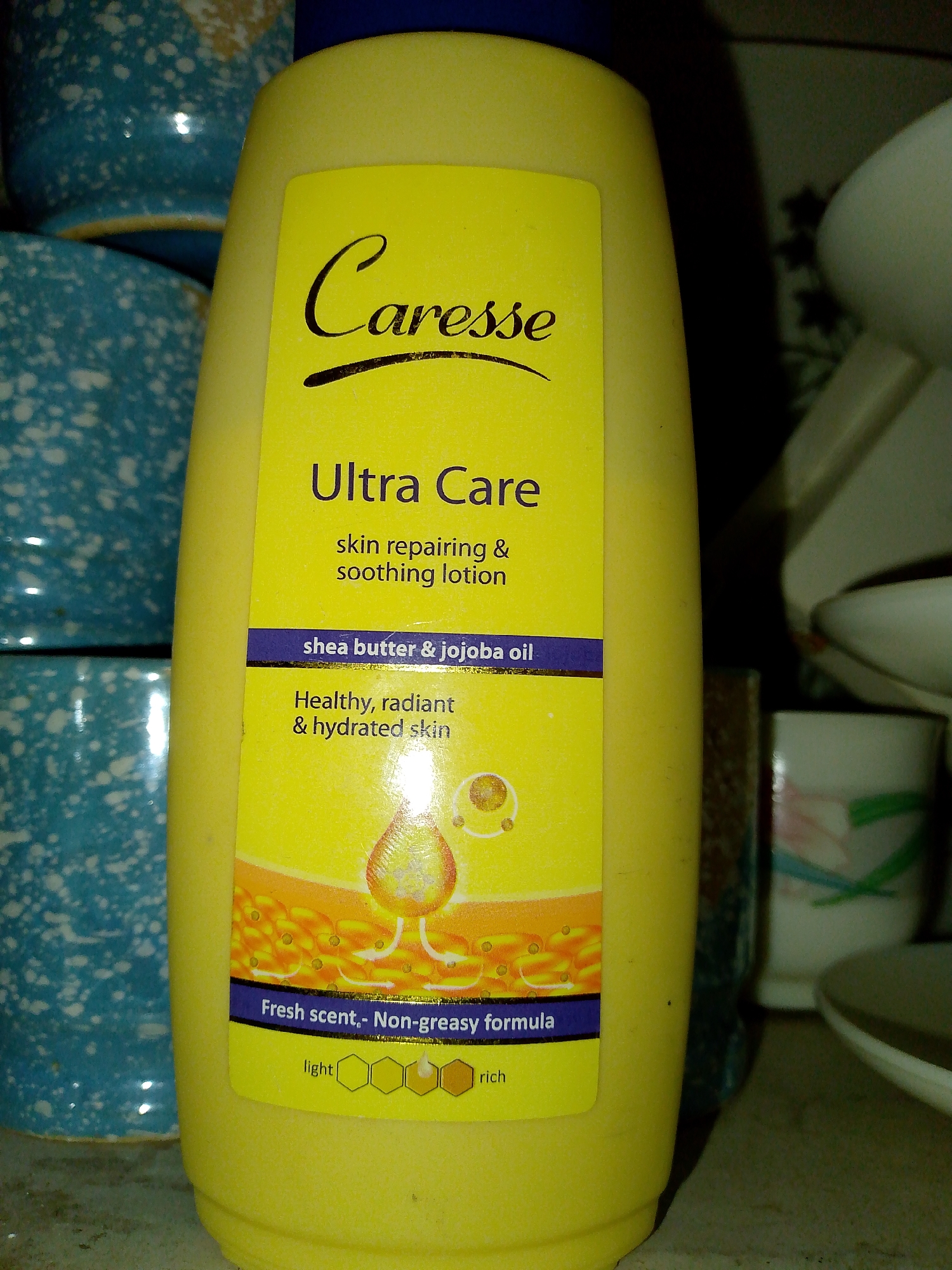 A bright yellow bottle of Caresse Ultra Care skin repairing and soothing lotion is positioned in the foreground. The label is yellow and features black text. The product contains shea butter and jojoba oil, and is described as providing healthy, radiant, and hydrated skin. It also boasts a fresh, non-greasy formula. At the bottom of the label, a gradient scale indicates the lotion's richness, from "light" to "rich." The bottle is surrounded by other objects, including blue and white speckled pottery in the background on the left, and white dishes or containers on the right. The lighting in the scene appears to be dim.