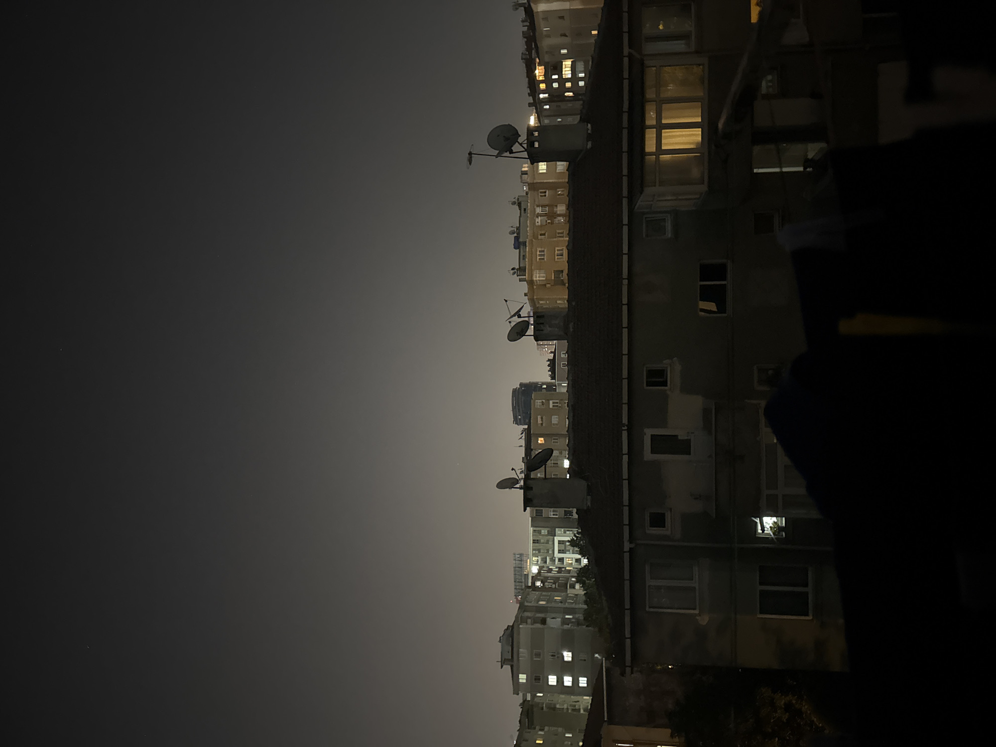 The image captures a nighttime cityscape in Gungoren, Turkey. The foreground is dominated by the dark silhouette of a building's roof and upper facade, obscuring much of the lower portion of the scene. Above this, a multi-story apartment building is visible, with several windows lit from within, casting a warm glow. Beyond this building, a cluster of taller apartment buildings stretches across the horizon, their windows also illuminated. The sky is a deep, hazy gray, typical of urban night skies, with a few faint stars or distant lights visible. Satellite dishes are mounted on chimneys atop some of the buildings, indicating residential occupancy. The overall impression is of a densely populated urban environment at night.