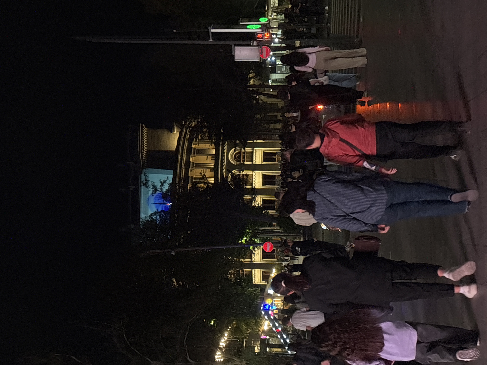 At night in Yerevan, Armenia, a crowd stands on a street near illuminated buildings. Traffic lights and signs are visible, while trees and street lights add to the atmosphere. Pedestrians are present.