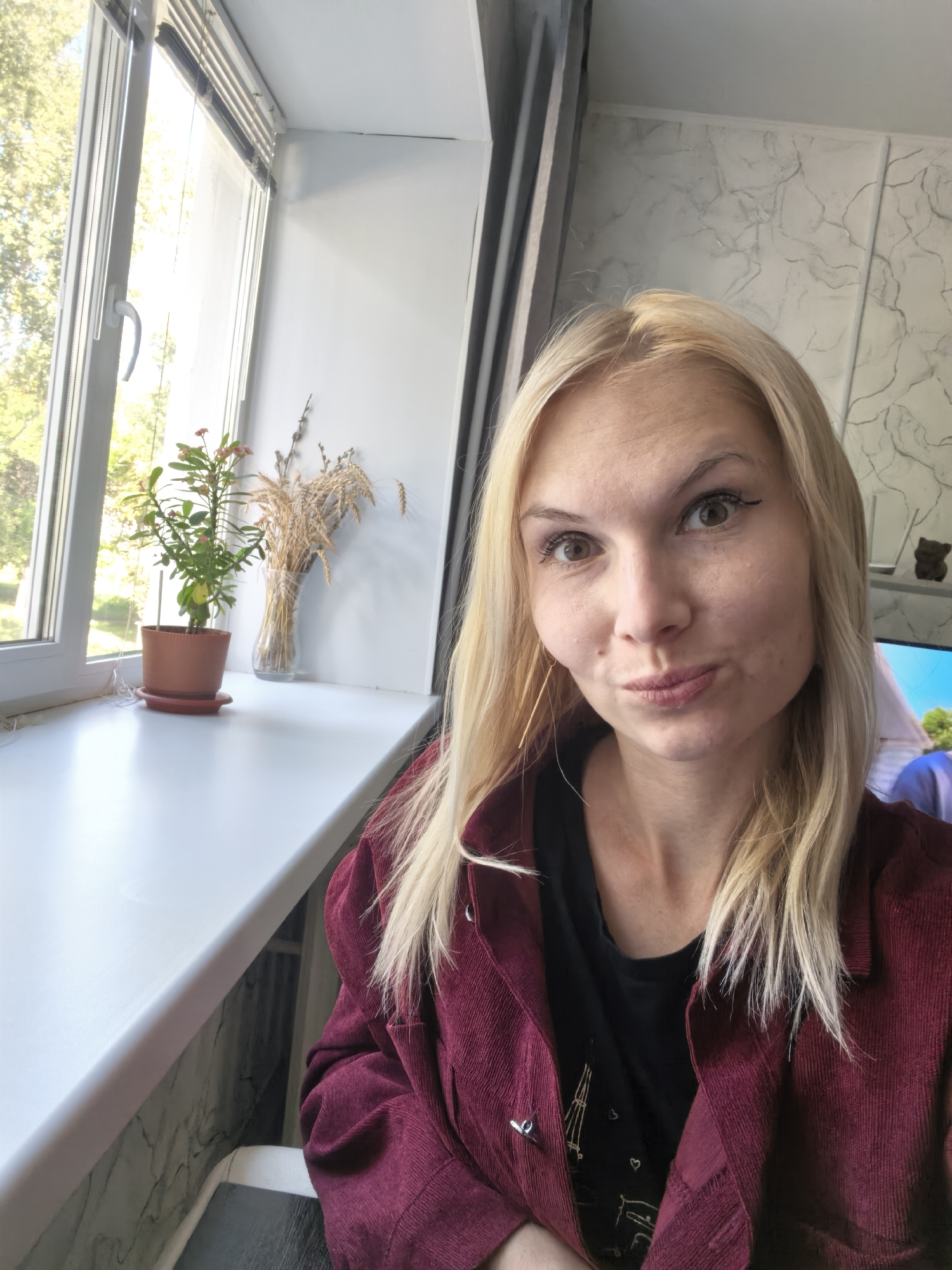 A blonde woman with light skin and light brown eyes is taking a selfie indoors, likely in her home. She is wearing a dark maroon corduroy jacket over a black t-shirt, and has on long, dangling earrings. Her hair is blonde and wavy, and she is wearing makeup, including eyeliner. She is puckering her lips and looking at the camera.
Behind her, to the left, is a window with blinds. Through the window, green trees and sunlight are visible, suggesting it is daytime and possibly spring or summer. On the windowsill, there is a small potted plant with green leaves and some pink flowers, and a glass vase filled with dried wheat stalks.
To the right of the woman, the wall has a textured, marbled pattern. A shelf with a small bear figurine and a television screen displaying an image of people and a landscape are visible in the background. The scene appears to be set in a residential interior. The location is not specified in the image or surrounding data.