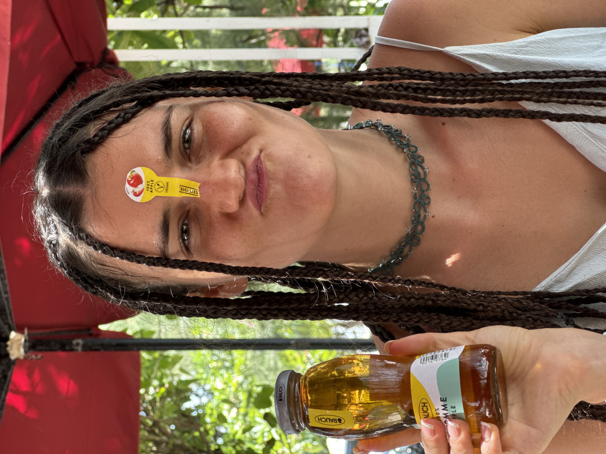 A young woman with dark braided hair is holding a glass bottle of apple juice. She is wearing a light-colored tank top and a green beaded necklace. A yellow sticker with an image of apples and the text "100% APPLE VEGAN" is affixed to her forehead. She is making a pouting face. The background shows greenery and red fabric, suggestive of an outdoor setting, possibly a restaurant with an umbrella. The woman's skin is lightly tanned, and she has freckles. The bottle is partially filled with a golden-colored liquid. The overall impression is a casual, outdoor scene. There is no information or visual evidence to suggest the location is in Phan Thiết, Vietnam.