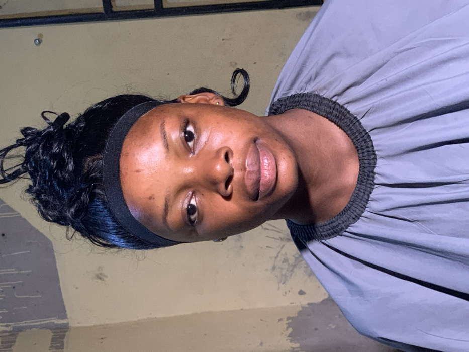 The scene depicts a young adult female, appearing to be of African descent, captured in a medium close-up shot indoors in Garaku, Nigeria. The specific location within Garaku, such as a street or building type, is not discernible from the visual evidence. She is facing directly forward, looking towards the camera. Her hair is styled in a dark, curly updo, secured by a black headband across her forehead. A singular dark curl hangs loosely behind her right ear. She is wearing a grey stylist's cape or similar protective garment, which features a dark, elasticized band around the neckline and visible pleats. The background consists of an interior wall painted in a faded light yellow or beige, showing areas of discoloration, possible paint drips, and plaster irregularities. A dark vertical frame, consistent with a window or door jamb, is visible on the right side of the image. No other individuals or significant objects are present within the frame.