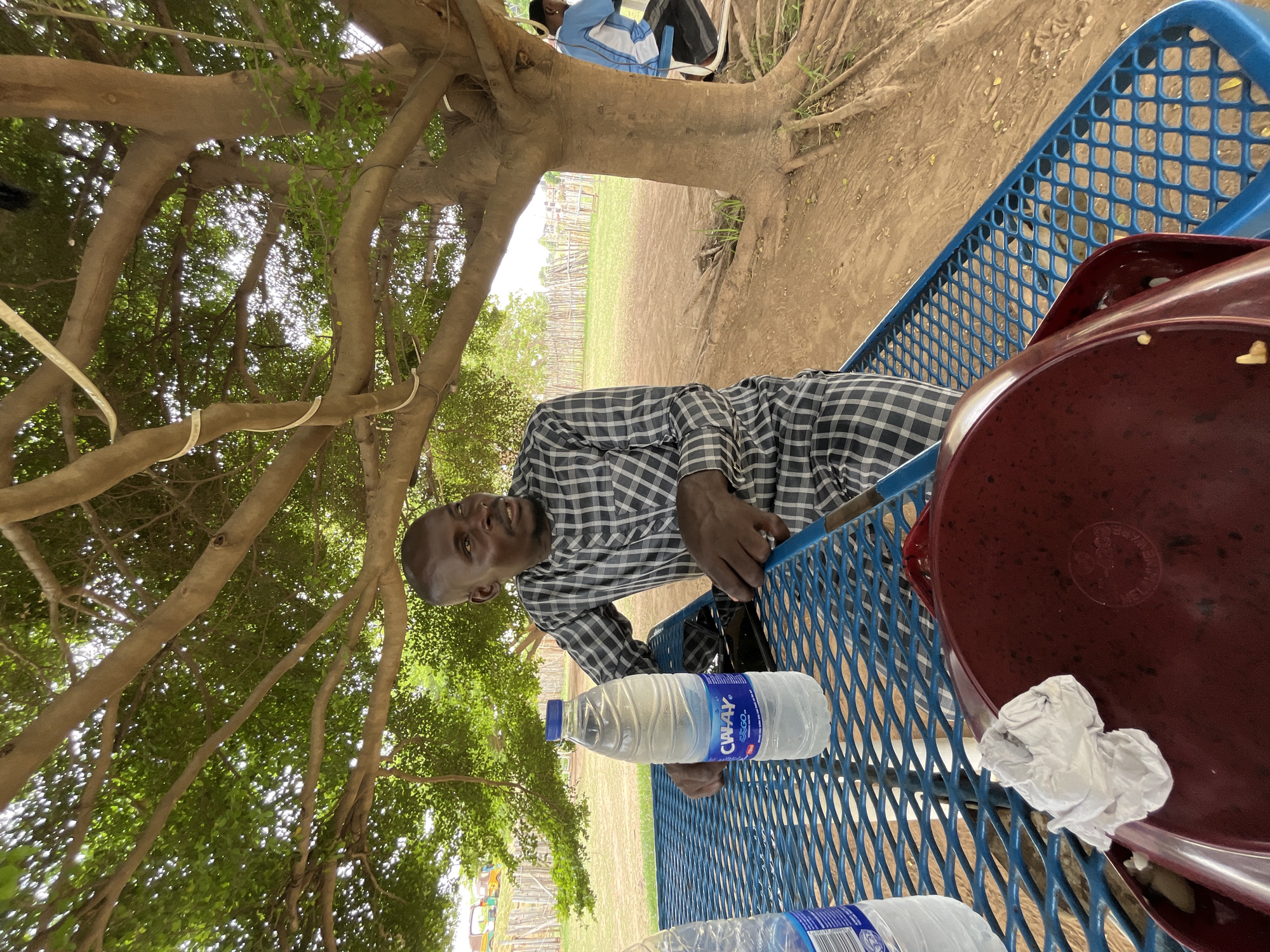 The image shows a man sitting at a blue picnic table under a large tree in an outdoor setting, likely in Galadimawa, Nigeria. The man is wearing a plaid shirt and is positioned at the table, where there are also bottles of water and a maroon food container with a crumpled white paper on top. The picnic table is situated on a dirt ground surface under the shade of the tree, which has expansive branches and green foliage. In the background, there is a field or open space enclosed by a fence. Another individual is seated in the background next to the tree's trunk.