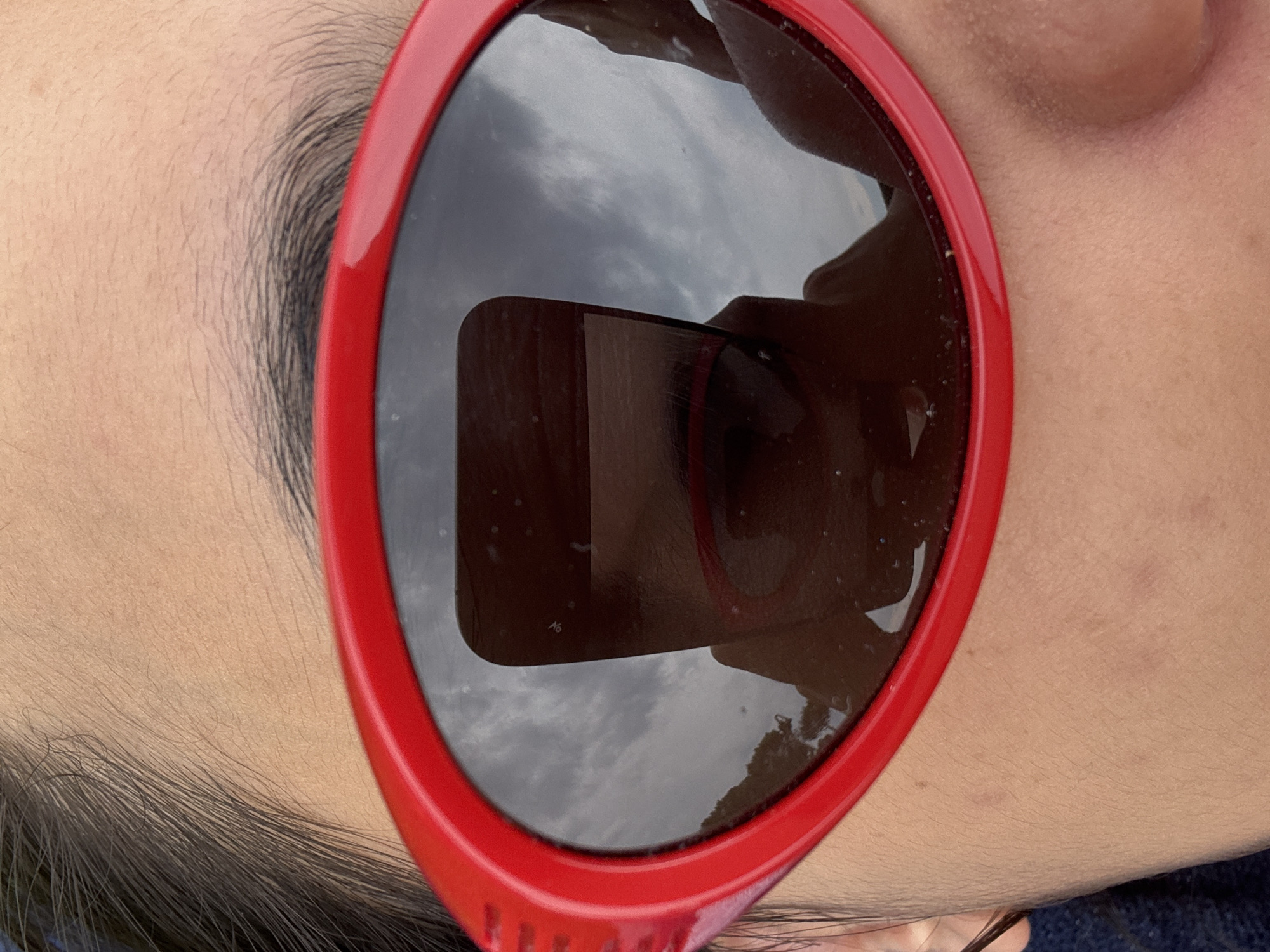 The image is a close-up, high-angle shot of a person wearing bright red, oversized sunglasses. The sunglasses have dark, reflective lenses that capture a reflection of a person holding a smartphone, presumably taking the photo. The reflection also shows a cloudy sky and a silhouette of trees. The person's skin, visible above and below the sunglasses, appears to be light-toned and slightly sun-kissed, with some minor imperfections. The background is out of focus, but the overall impression is an outdoor setting, possibly during the daytime, given the reflection of the sky. There is no specific indicator of the location being El Escorial, Spain, in the image itself.