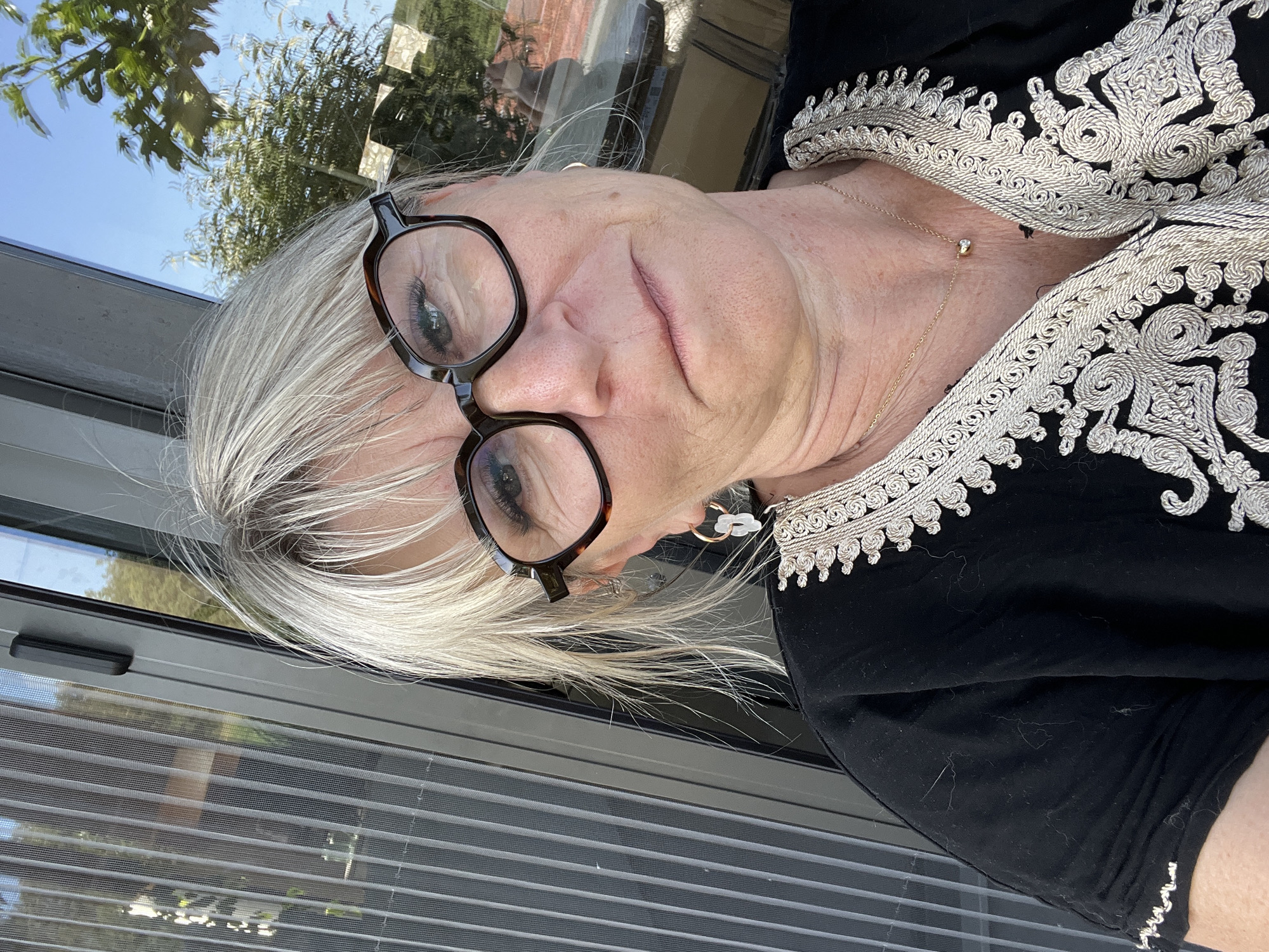 A woman is taking a selfie outdoors. She is wearing a black garment with white embroidery and glasses with tortoiseshell frames. She has blonde hair and is wearing a necklace and earrings. She is looking at the camera with a slight frown. In the background, there is a window and some greenery, suggesting she is near a building. The image is a close-up of the woman's face and upper body. There is no information about the location in Chambœuf, France.