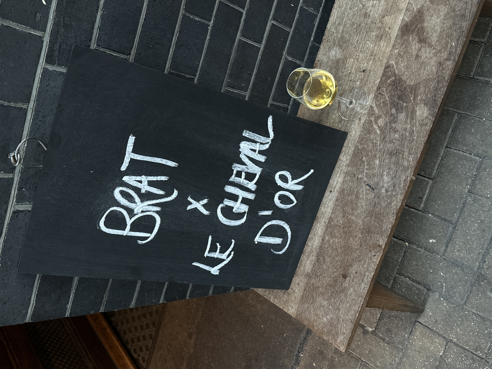 The image shows a close-up view of a blackboard sign resting on a wooden table, with a glass of white wine beside it. The sign displays the text "Brat x Le Cheval D'Or" in white chalk. The background consists of a dark brick wall and a paved sidewalk. The scene is likely located in an outdoor area, possibly a restaurant or bar patio. The style of the sign and the presence of wine suggest an establishment located in London, United Kingdom.
