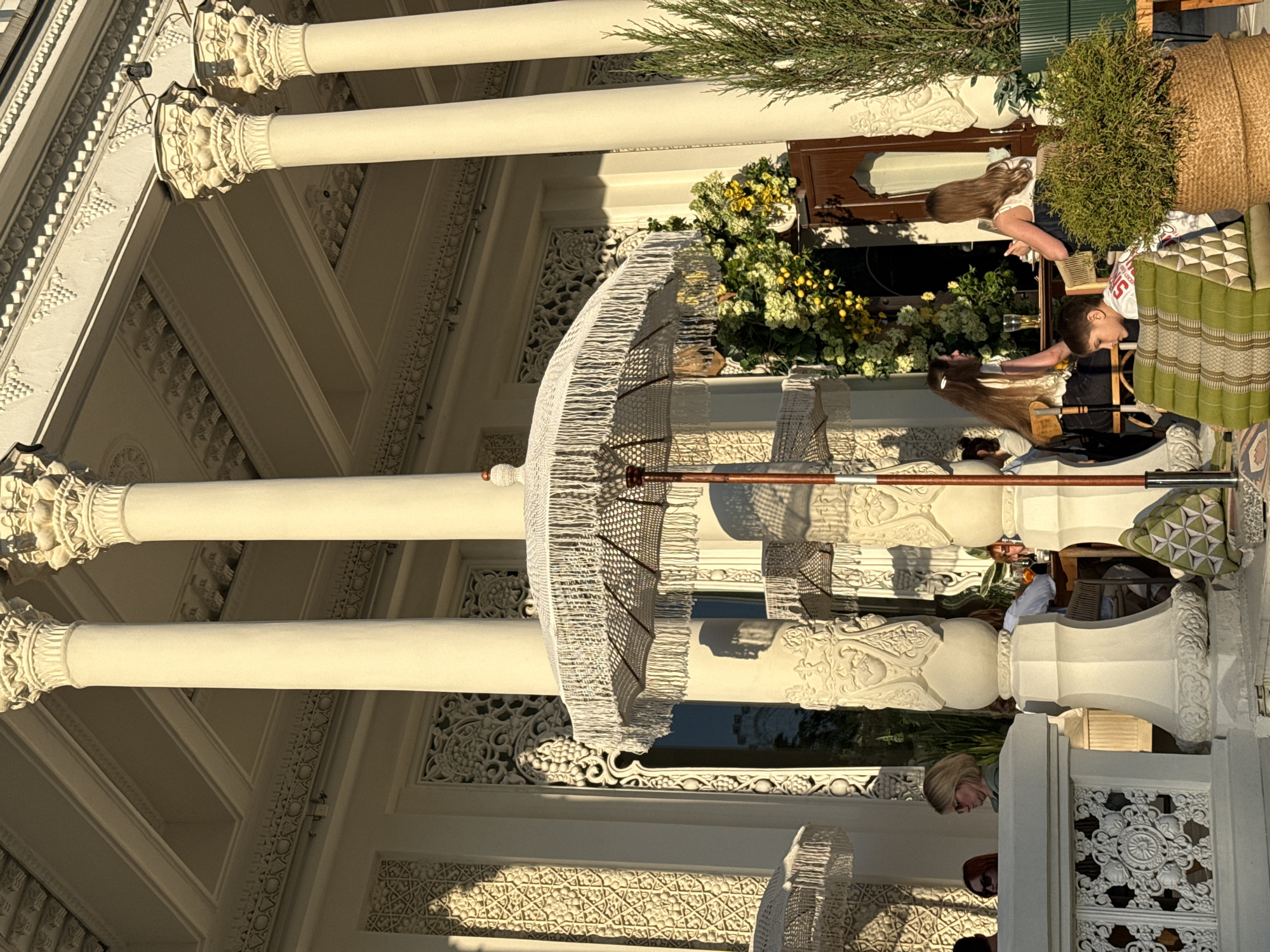 The image captures a scene on the veranda of the Pashkov House in Moscow, Russia. The veranda is adorned with ornate white columns and decorative carvings on the facade. A white fringed parasol provides shade. People are present, including two individuals seated at a table. Plants and flowers, along with furniture, contribute to the ambiance of the space.