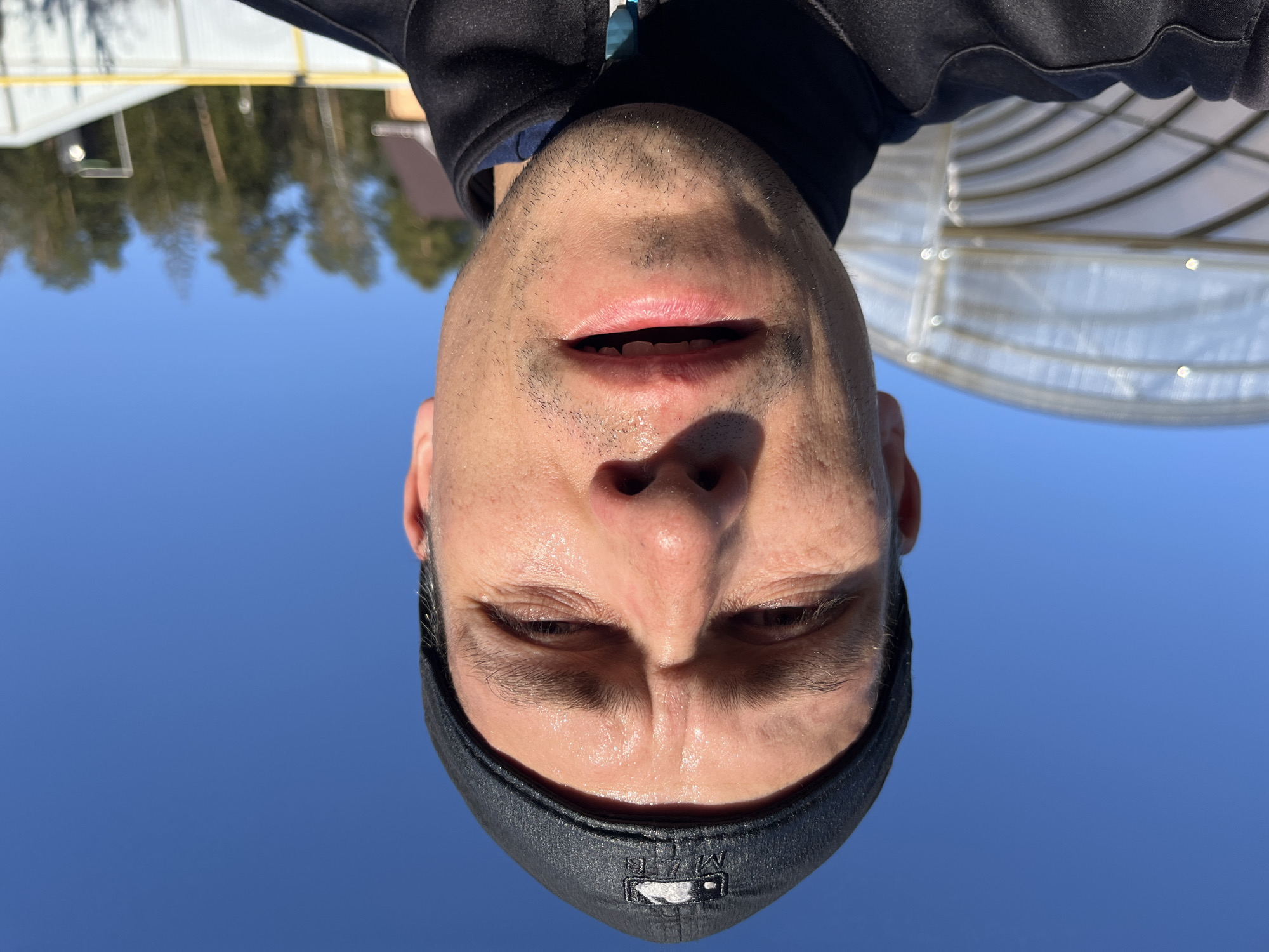 The image is an upside-down selfie of a man wearing a black baseball cap. The background shows a clear blue sky, a blurry treeline, and a semi-circular metal structure with translucent panels. The location is likely in or around the city of Chaikovsky, Russia.