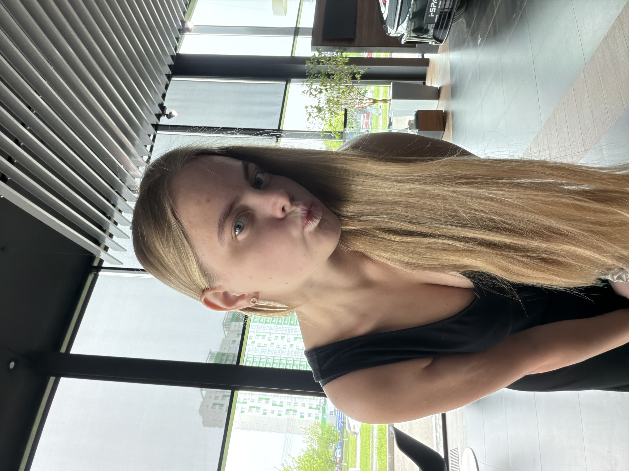 A blonde woman with fair skin is posing for a selfie indoors. She has straight blonde hair and is wearing a black dress. Her lips are pursed in a kissing expression. The background includes a window showing buildings with a lot of green. Inside the building, there's a modern ceiling structure and a car dealership. The location appears to be in the city of Izhevsk, Russia, as indicated by the signs outside the window.
