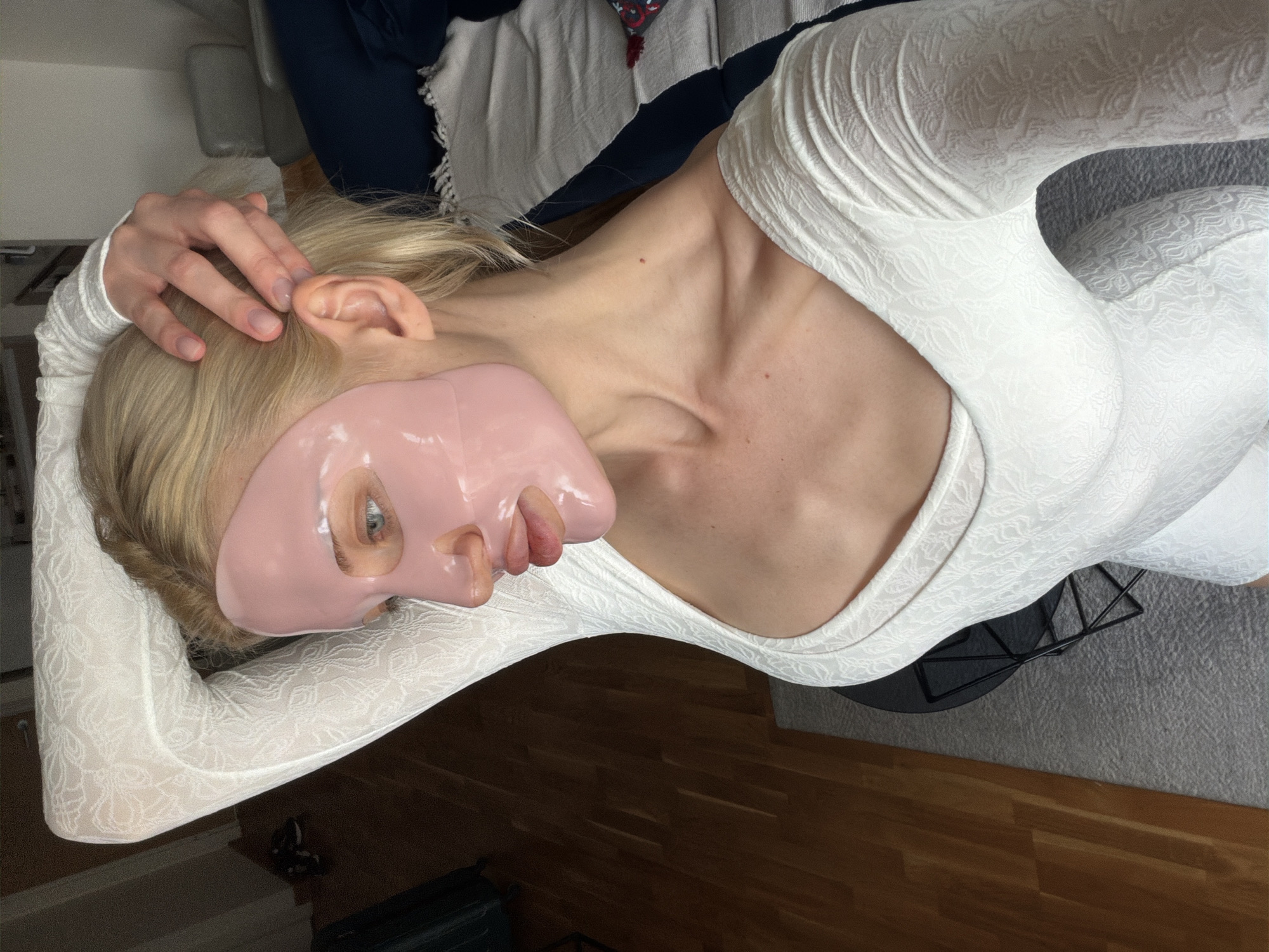 A blonde woman with a pink face mask is lying down, dressed in a white, long-sleeved, textured top. Her right hand is resting on her head. She is resting in a room with wood flooring and a gray rug. A small black table is next to her. In the background, there is a bed with a white blanket. The location is likely indoors, but not definitively Belgrade, Serbia based on the current information.