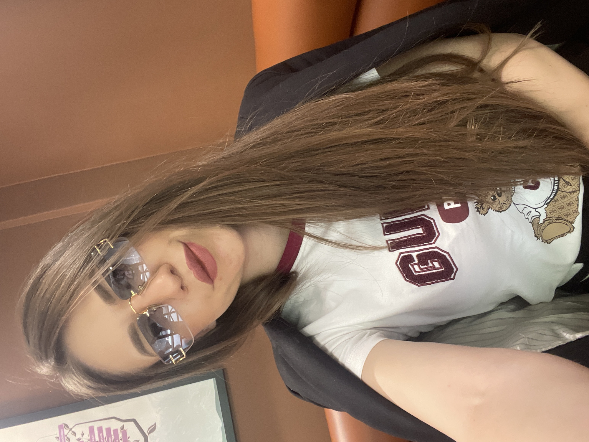A woman is taking a selfie indoors. She has long, brown hair, wears sunglasses, and a white t-shirt with the word "GUU" in maroon letters. A black blazer is draped over her. The background features a brown wall and a piece of framed art. The location is not discernible from the image.