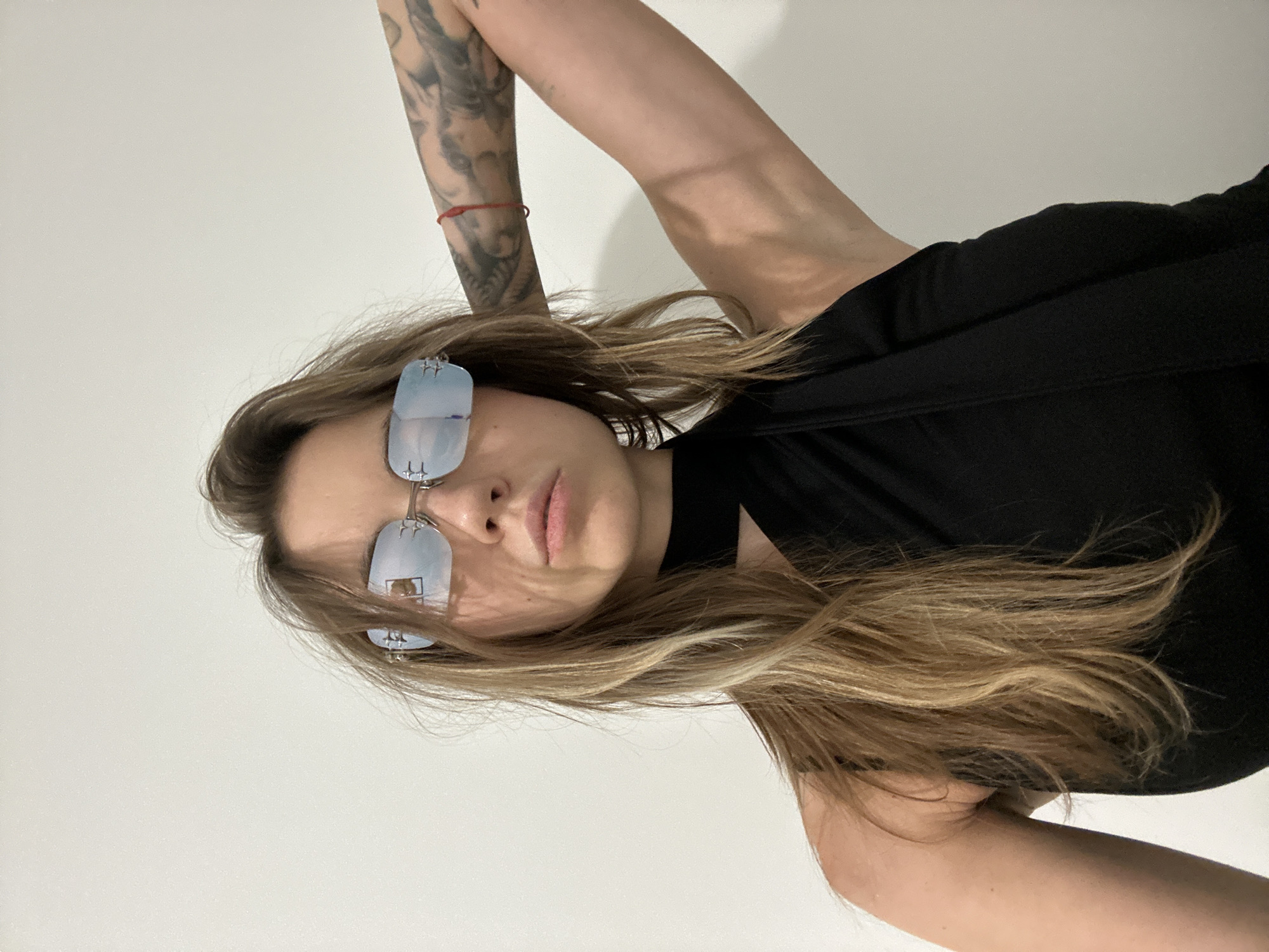 A young woman with long, blonde hair, wearing rimless, light blue tinted sunglasses and a black top, poses with her right arm raised behind her head. She has a red string bracelet on her left wrist and tattoos on her right arm. The background is a plain white wall, suggesting an indoor setting. No specific location in Moscow, Russia, can be determined from the image.