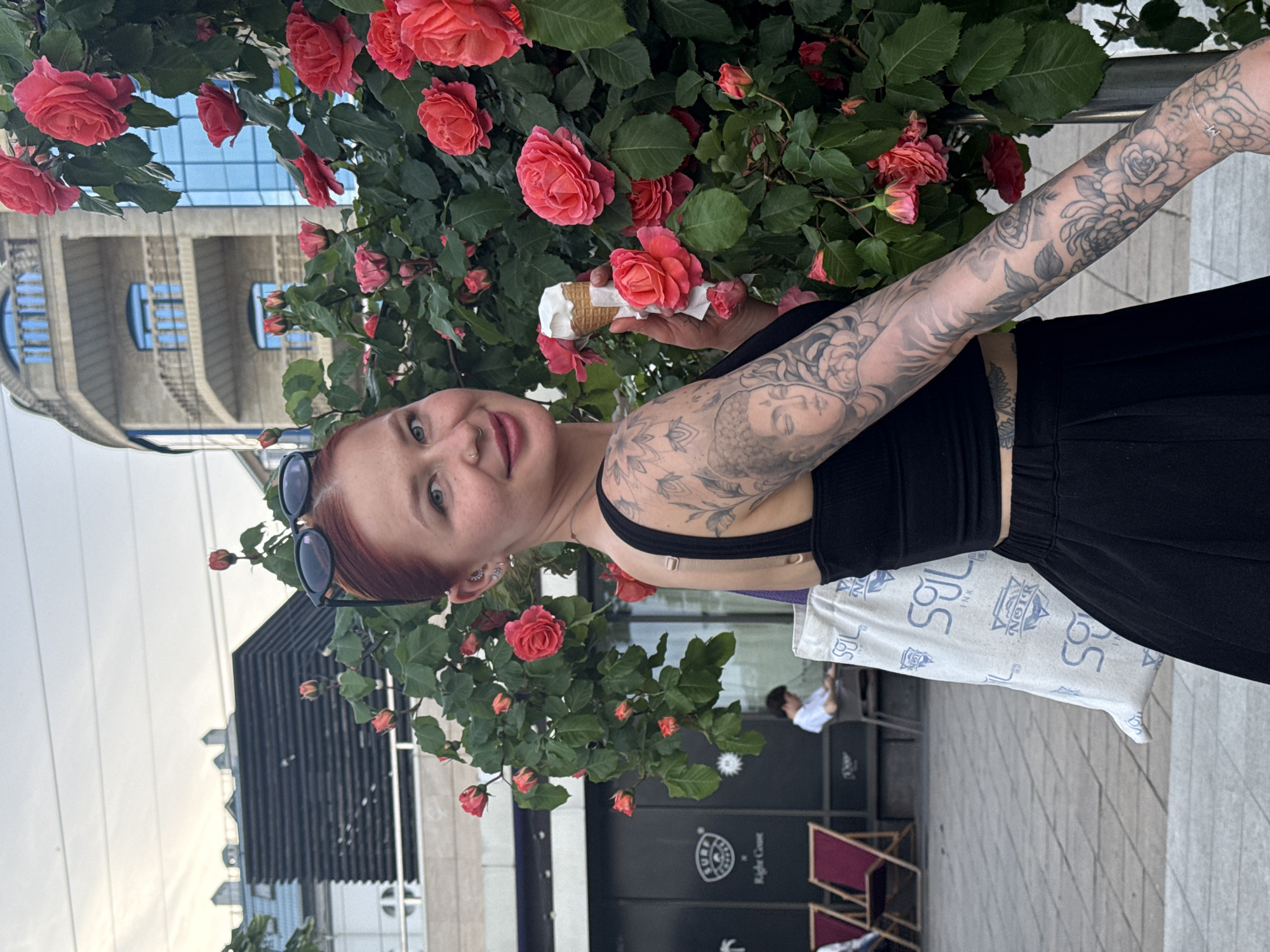 A young woman with fair skin, red hair, and tattoos on her arm is standing outside near a large bush of pink roses. She is holding an ice cream cone in her left hand. She is wearing a black crop top and black pants. A pair of sunglasses are perched on her head. In the background, there is a building with a curved balcony and a modern black building. A store sign with Cyrillic letters is visible on the black building. There's a white printed bag and some chairs nearby. People can be seen sitting at tables in the background. The scene is outdoors, likely in Rostov-on-Don, Russia based on the Cyrillic writing.