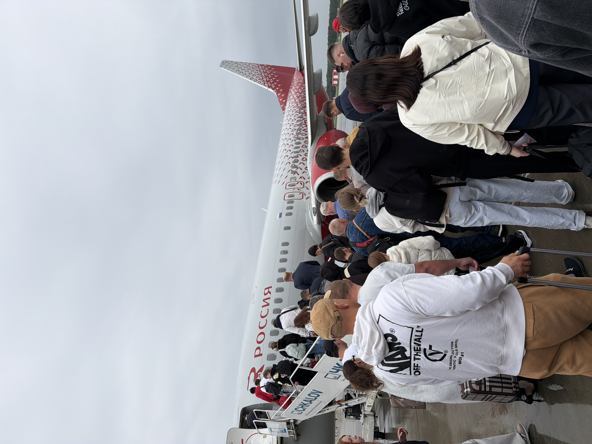 The image depicts passengers boarding a Russia Airlines plane at Chkalov Airport in Nizhny Novgorod, Russia. A group of people, dressed in casual attire and carrying luggage, are ascending a set of stairs to enter the aircraft. The plane, a Boeing 737, has the word "РОССИЯ" (Russia) written in red on its fuselage, and its tail features a distinctive red and white geometric pattern. The sky is overcast, suggesting a cloudy day. The ground appears wet, possibly from recent rain.