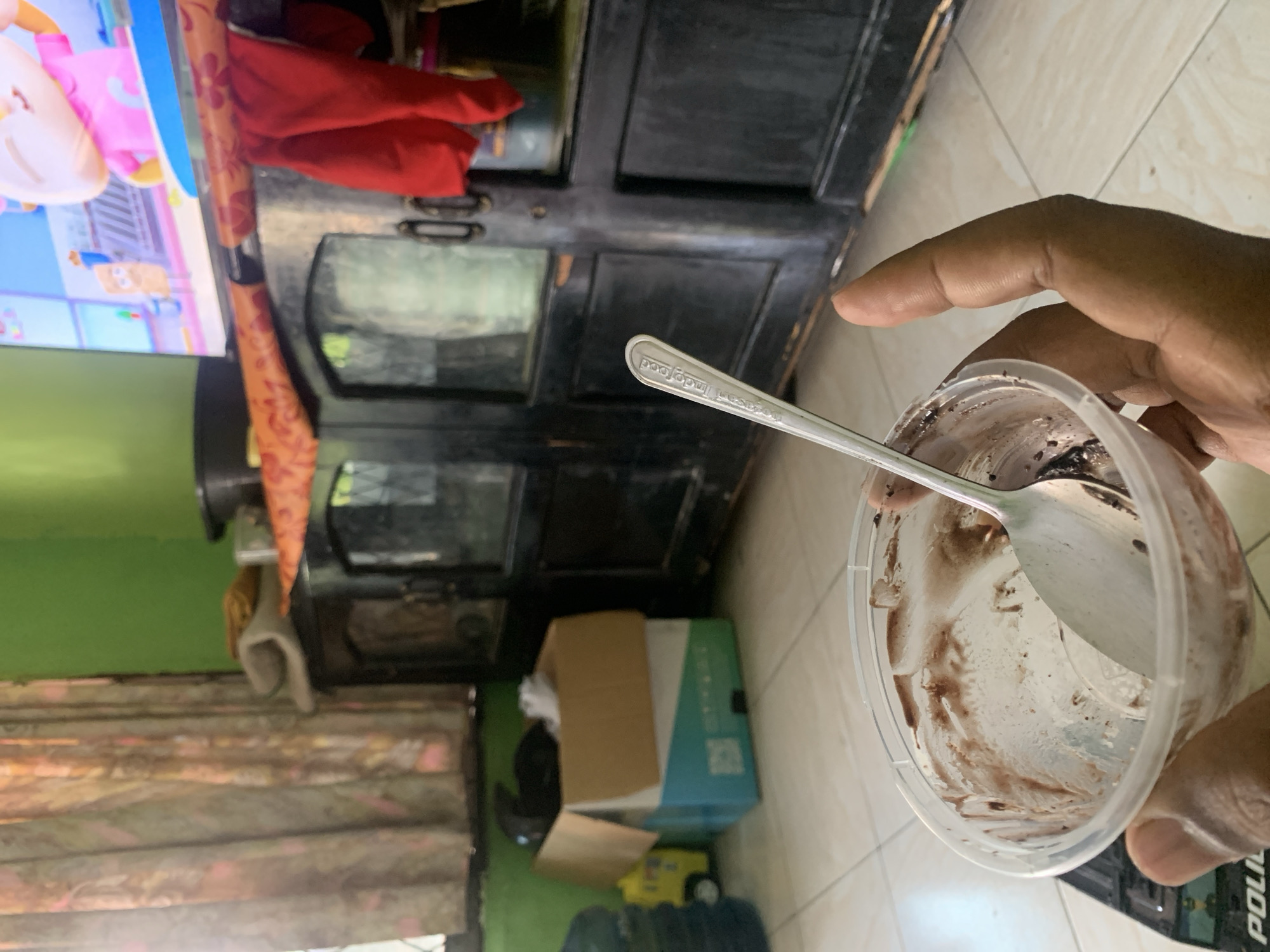A person is holding a nearly empty plastic ice cream container with a spoon inside, with chocolate ice cream residue visible on the sides and spoon. The container is clear plastic, and the spoon appears to be made of metal, with the word "Indofood" embossed on its handle. In the background, there is a dark wooden cabinet with glass doors, a television displaying a cartoon, red cloth draped over it, and curtains with a floral pattern. To the left, there's a cardboard box and a blue and white box. The floor is tiled. The scene appears to be indoors, in what could be a living room or dining area. The presence of a television and furniture suggests a residential setting, likely in Palembang, Indonesia, given the "Indofood" branding on the spoon, which is a prominent Indonesian food company.