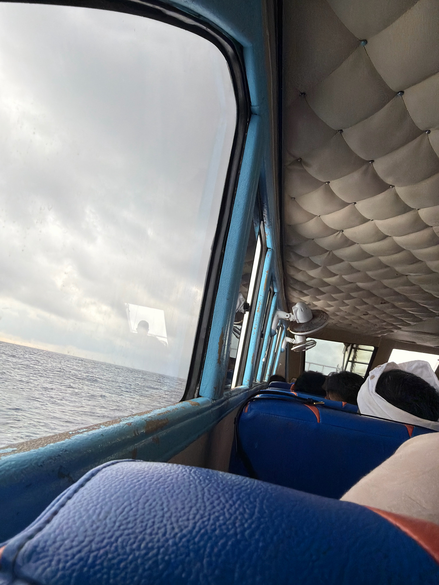 The image shows the interior of a passenger boat, likely in Indonesia. Through a large window, the sea and a cloudy sky are visible. Inside, the ceiling is padded with a diamond-patterned material, and a small fan is mounted on the ceiling. Blue and orange upholstered seats are arranged in rows, with several passengers seated. A person wearing a white head covering is visible in the foreground on the right. The overall impression is one of travel on a mode of transport at sea.