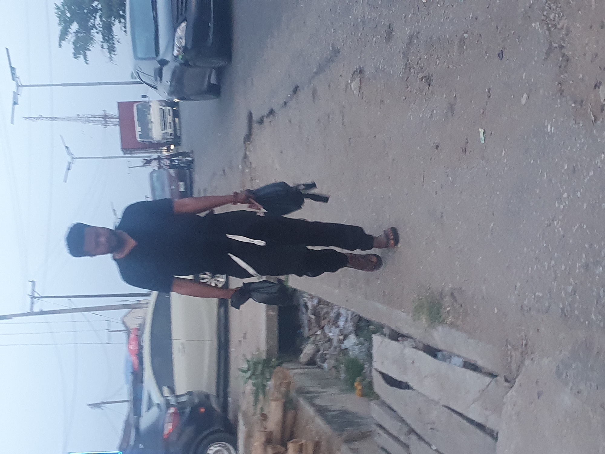 The image shows a man walking along a street in Lokoja, Nigeria. He is wearing dark clothing and sandals, and carrying a bag. The street is lined with parked cars, and a truck is visible in the distance. Power lines and transmission towers are also visible in the background. The environment appears to be an urban area. The weather is overcast or foggy.