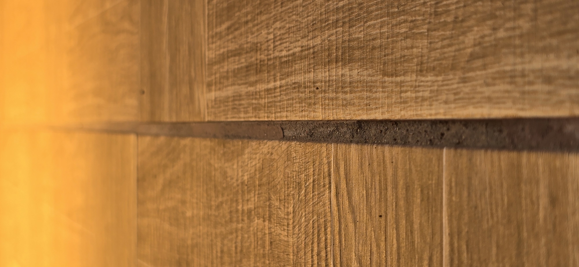 The image displays a close-up, vertical view of a weathered wooden floor with a prominent, dark grout line running down the center. The wood planks are oriented horizontally, with visible grain patterns and some surface imperfections, suggesting age and use. The lighting creates a warm, golden hue across the scene, with a gradient from brighter at the top to slightly darker towards the bottom. The grout line appears rough and uneven, filled with what looks like a dark, granular material. There are no people or discernible actions depicted. The scene is devoid of any specific context that would tie it to Urupskiy, Russia, or any other urban environment.