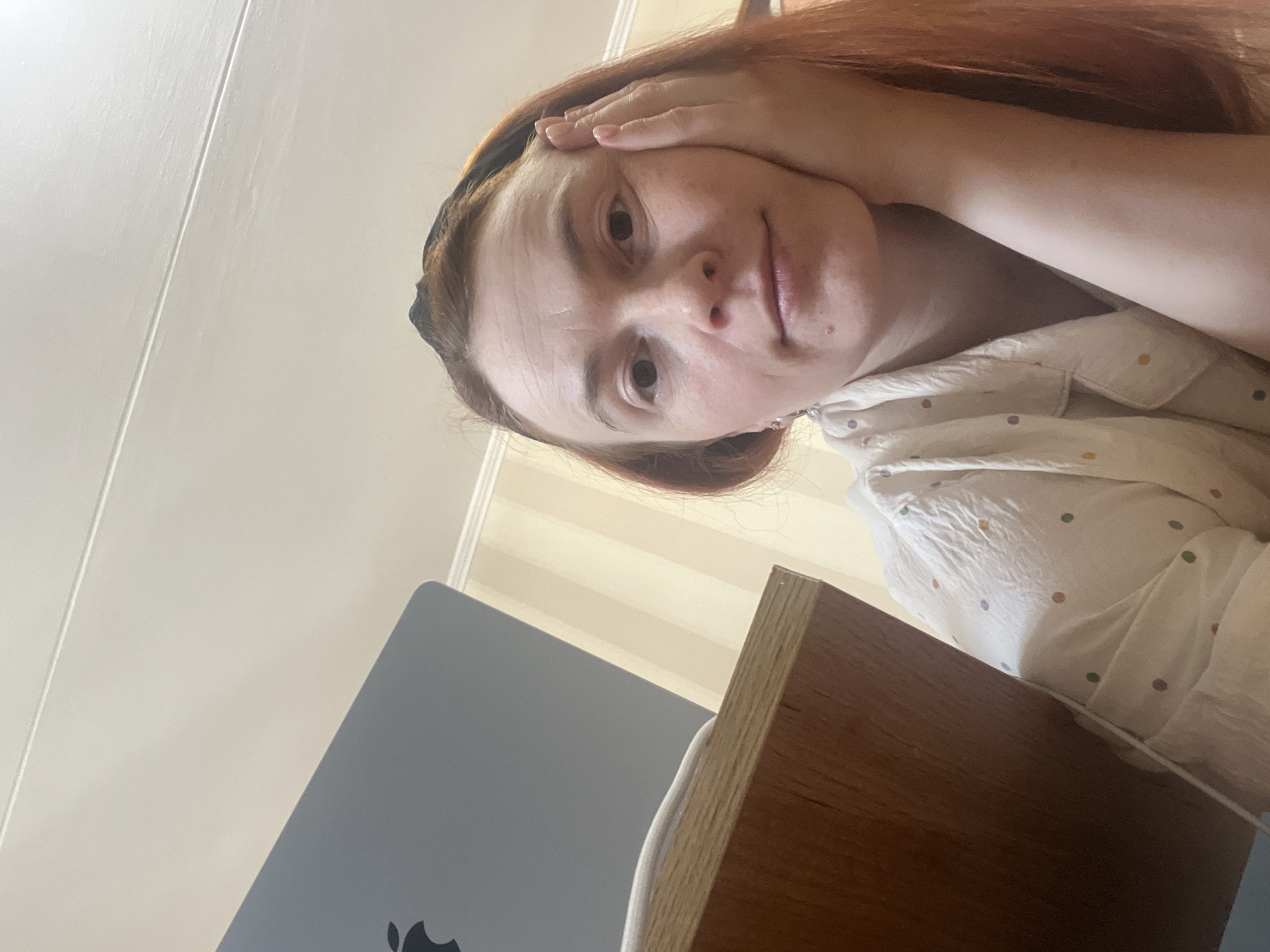 A selfie taken from a high angle shows a woman with red hair and a headband. She is wearing a white, collared shirt with multicolored polka dots. Her left hand is resting on her forehead. A gray laptop with an Apple logo is visible in the lower left corner of the frame, partially obscured by a wooden desk. The background includes a light-colored wall with vertical stripes. The location is Chaikovsky, Russia.