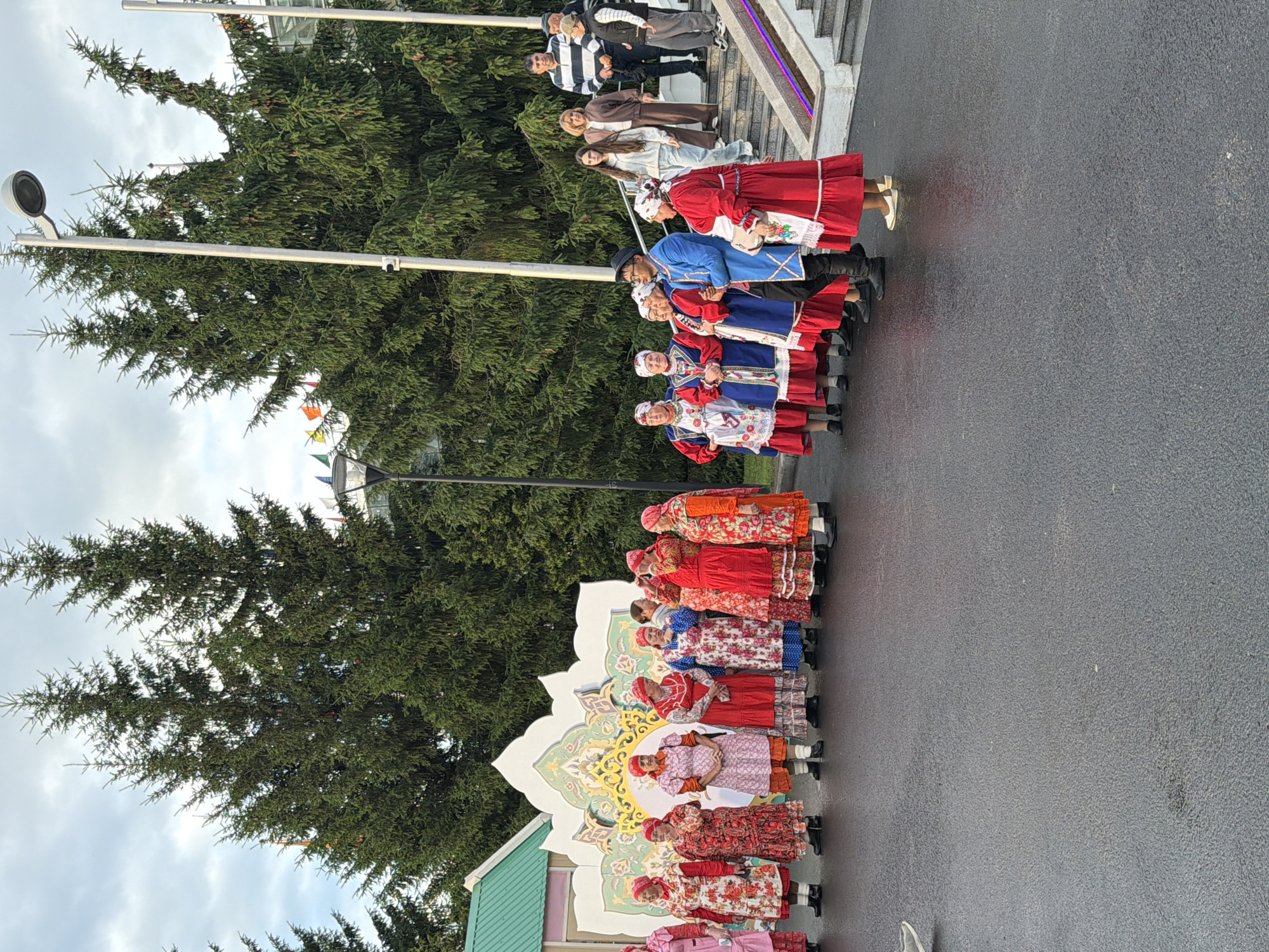 In an outdoor setting with a backdrop of large evergreen trees, a group of people, predominantly women dressed in traditional colorful folk costumes, are gathered. The women are standing in a line on a wet asphalt surface. They are wearing long, patterned dresses and headscarves. Some are interacting with each other, looking at the camera, or standing with their hands clasped. Behind them, a large, ornate decorative backdrop with floral motifs is visible. Further to the right, a set of stairs ascends, with several individuals dressed in modern clothing standing on it. A streetlamp is also present on the right side of the image. The overall atmosphere suggests a cultural event or performance.
