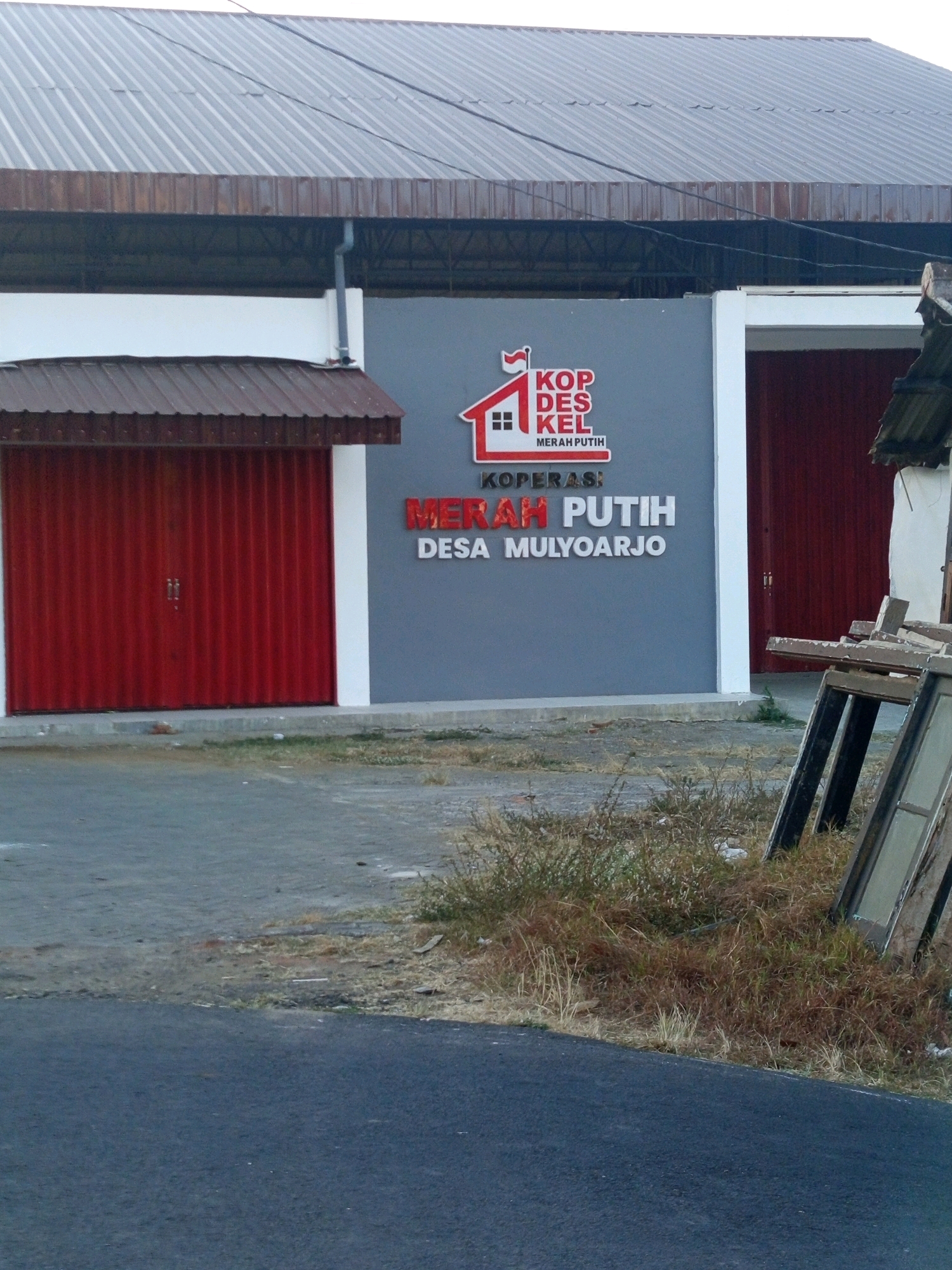 The image depicts the facade of a building with a prominent gray wall featuring a red and white logo and text in Indonesian. The logo is a stylized house with a red roof and a white outline, accompanied by the text "KOP DES KEL" in red and "MERAH PUTIH" in smaller red text below. Below this logo, larger red text reads "MERAH PUTIH" and beneath that, smaller white text reads "DESA MULYOARJO". To the left of the gray wall, there is a red corrugated metal roll-up door, partially open, with a brown overhang above it. To the right of the gray wall, another red corrugated metal roll-up door is visible, this one closed. The building has a corrugated metal roof that extends over the structures. The foreground shows an asphalt road and an area with dry grass and some discarded wooden frames and window panes. The scene suggests a commercial or community building in Desa Mulyorejo, Indonesia.