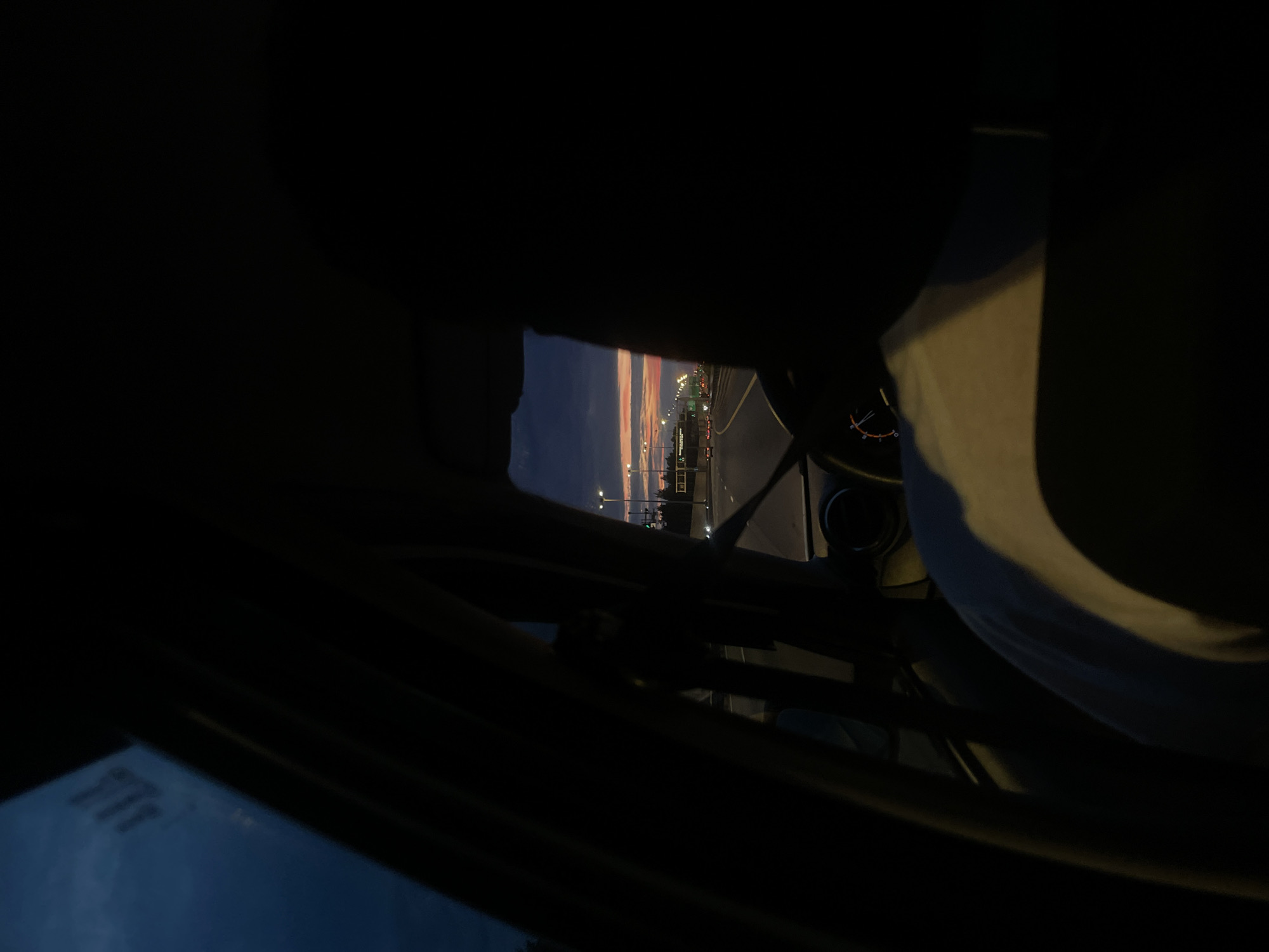The image depicts a view from the interior of a car, looking out towards a highway at dusk. The sky is a gradient of dark blue and orange, indicating either sunrise or sunset. Streetlights line the highway, and a few cars are visible in the distance. The foreground is dark, with the silhouette of a person's head and shoulder visible on the right side. Part of the car's dashboard and steering wheel are also in view on the left side. The image suggests a journey on a road near Pesochny, Russia, given the context provided.