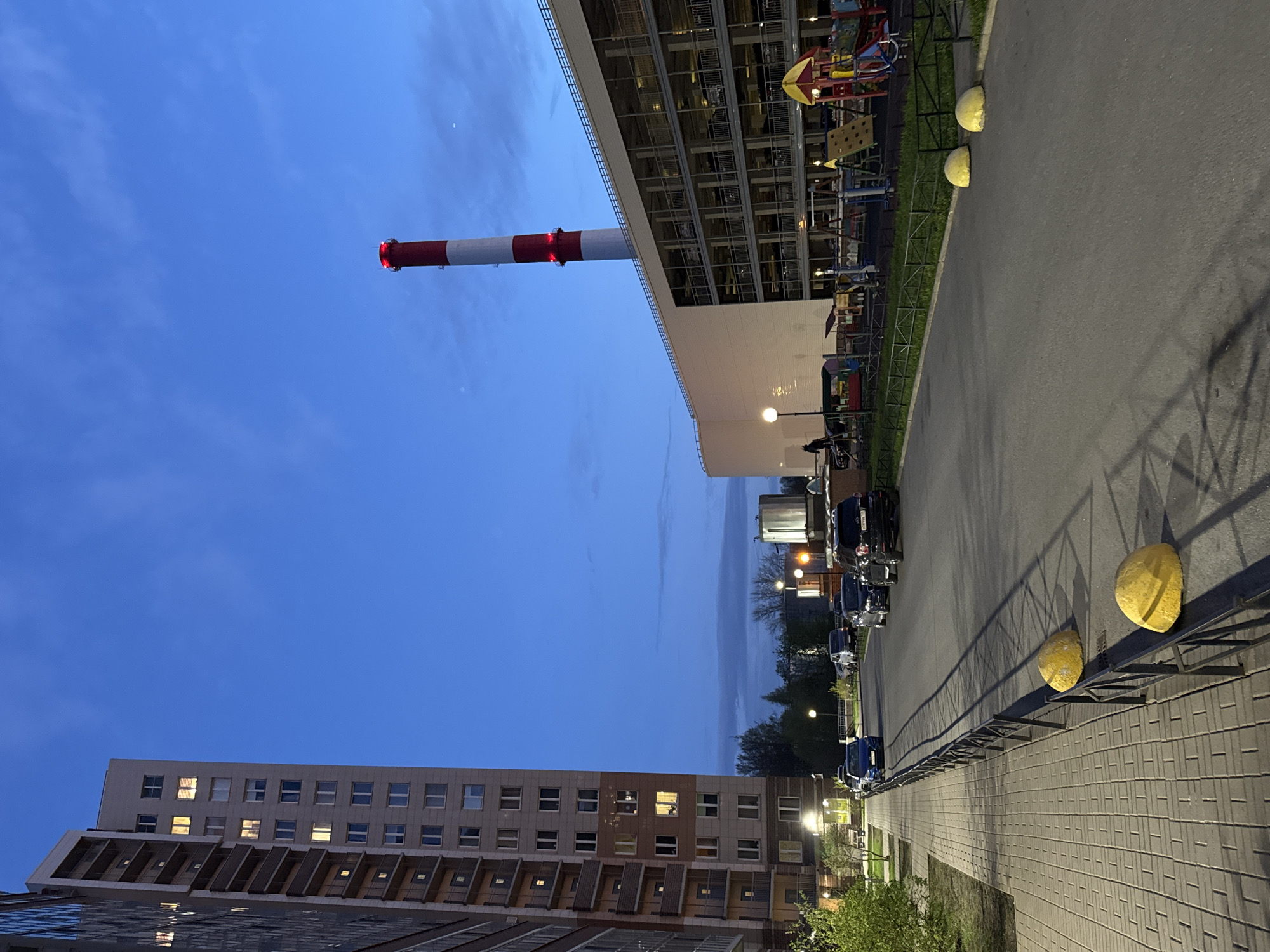 The image depicts an urban landscape in Saint Petersburg, Russia, during the evening hours. The sky is a gradient of blue, transitioning from a deep twilight hue to a lighter shade near the horizon. A tall industrial chimney with red and white stripes is a prominent feature, situated near a large, modern building with a multi-story parking structure visible on its upper levels, accompanied by a playground. Adjacent to the parking structure is a street lined with parked cars. The ground level features a paved walkway and a curb with spherical yellow traffic barriers. In the distance, another residential building with lighted windows is visible. No people are visible in the immediate scene. The overall setting appears to be a residential area near industrial or commercial structures.