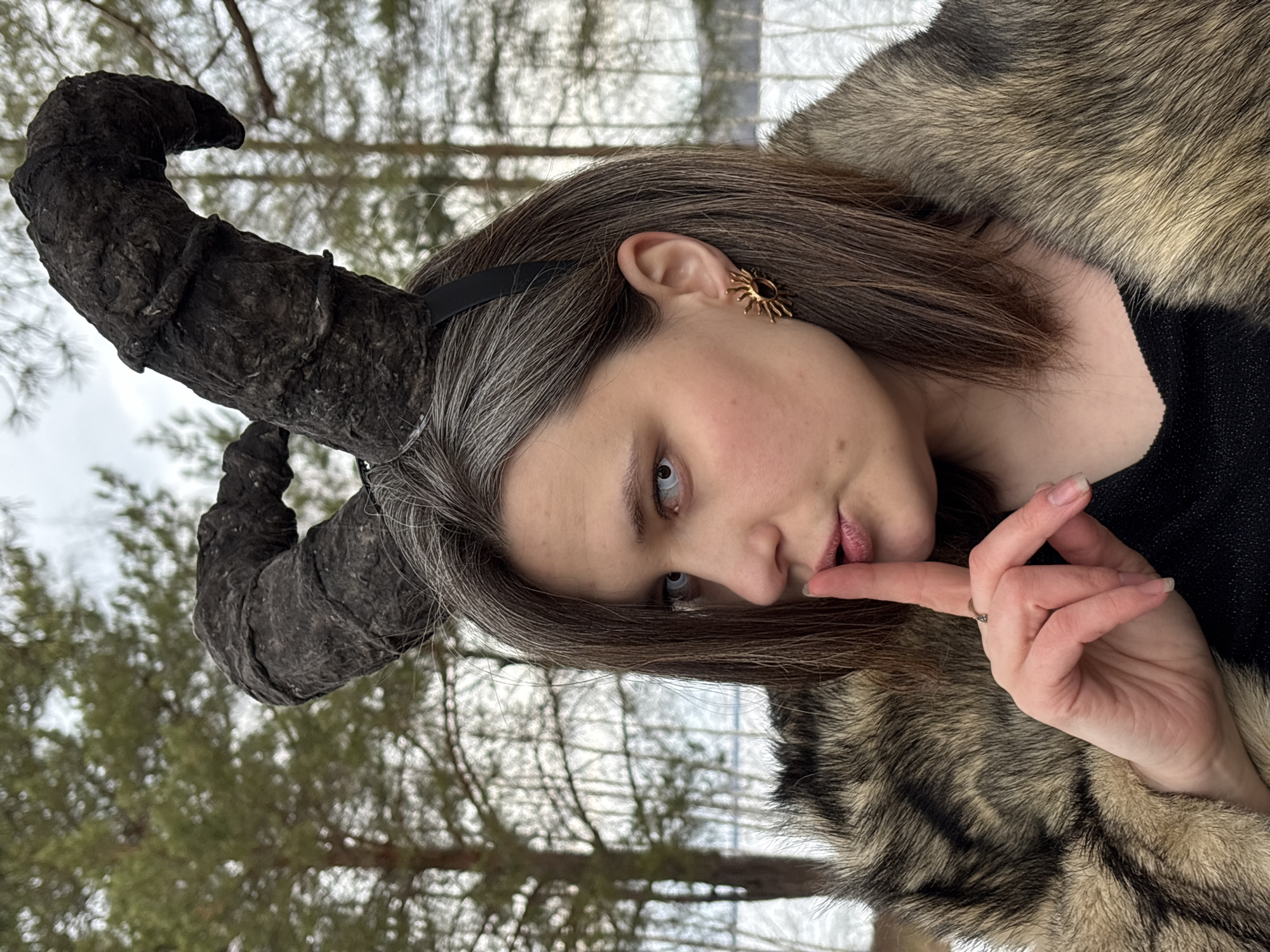 A woman with graying brown hair, fair skin, and light blue eyes is the focus of an outdoor photograph. She is wearing a black headband with large, textured black horns attached, and a fur-trimmed garment. Her right hand is raised, with her index finger touching her lips. She is wearing a gold-colored sun-shaped earring in her left ear. The background features snow-covered ground and bare trees, characteristic of a winter landscape. Although the prompt mentions Chaikovsky, Russia, there is no specific information in the image to confirm that location.