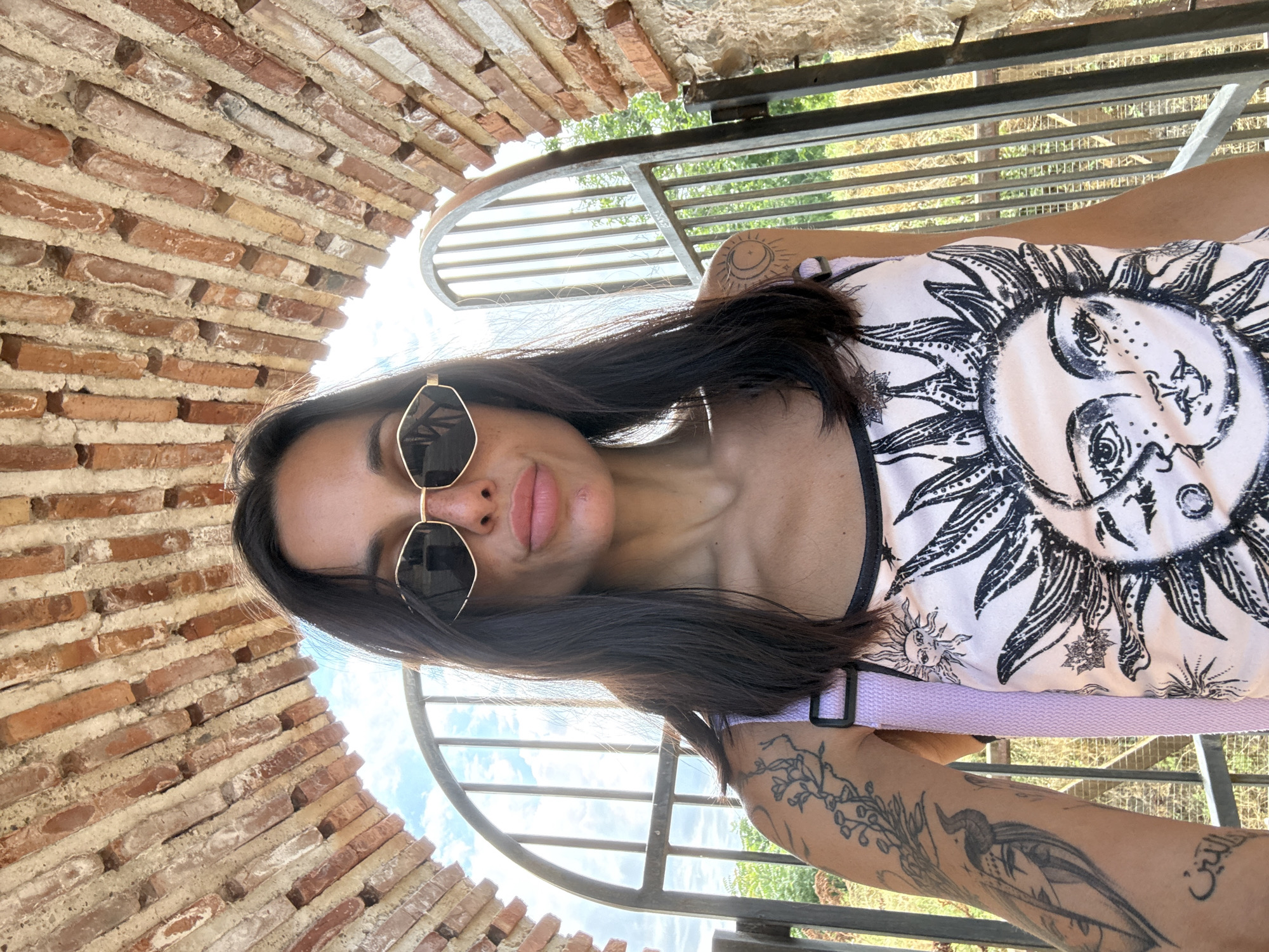 A woman with dark hair and sunglasses stands under a brick archway. She is wearing a light-colored tank top with a sun graphic and has tattoos on her arms. Behind her is a metal gate and an outdoor setting with greenery under a bright sky. The image appears to have been taken in the city of Belene, Bulgaria, possibly near the ruins of the monastery complex on the island of Persin.