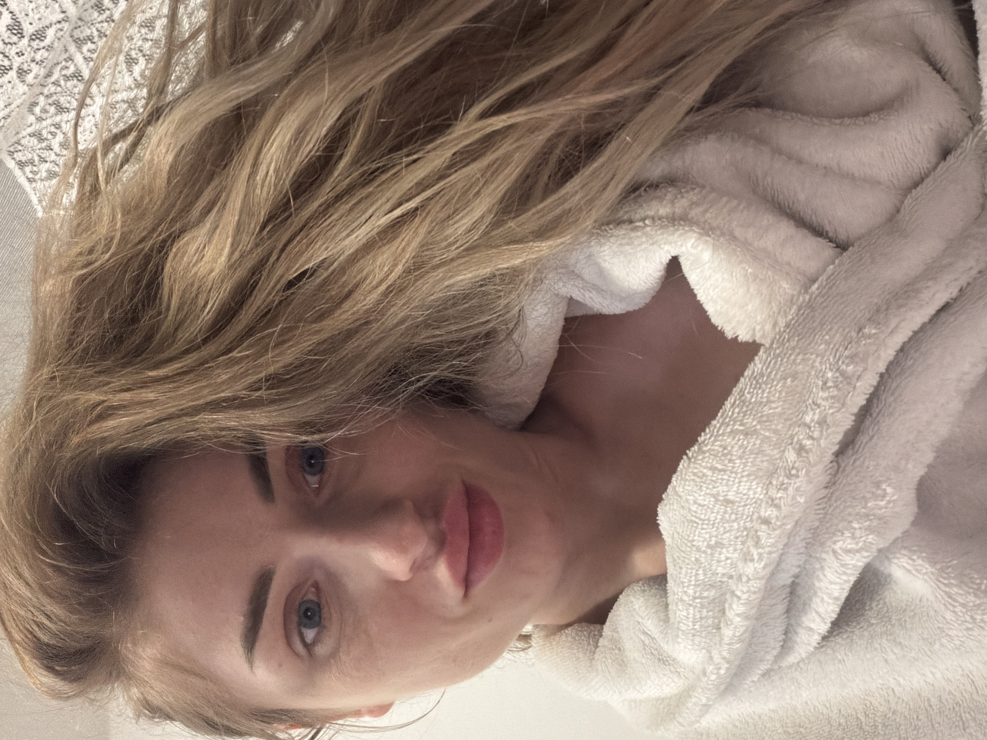 A medium shot portrait shows a blonde woman in a white bathrobe looking at the camera. Her hair is long and light brown, falling over her right shoulder. Her eyes are blue and her lips are pink and slightly pursed. A white lace fabric is visible in the upper right corner of the image. The background is plain white.