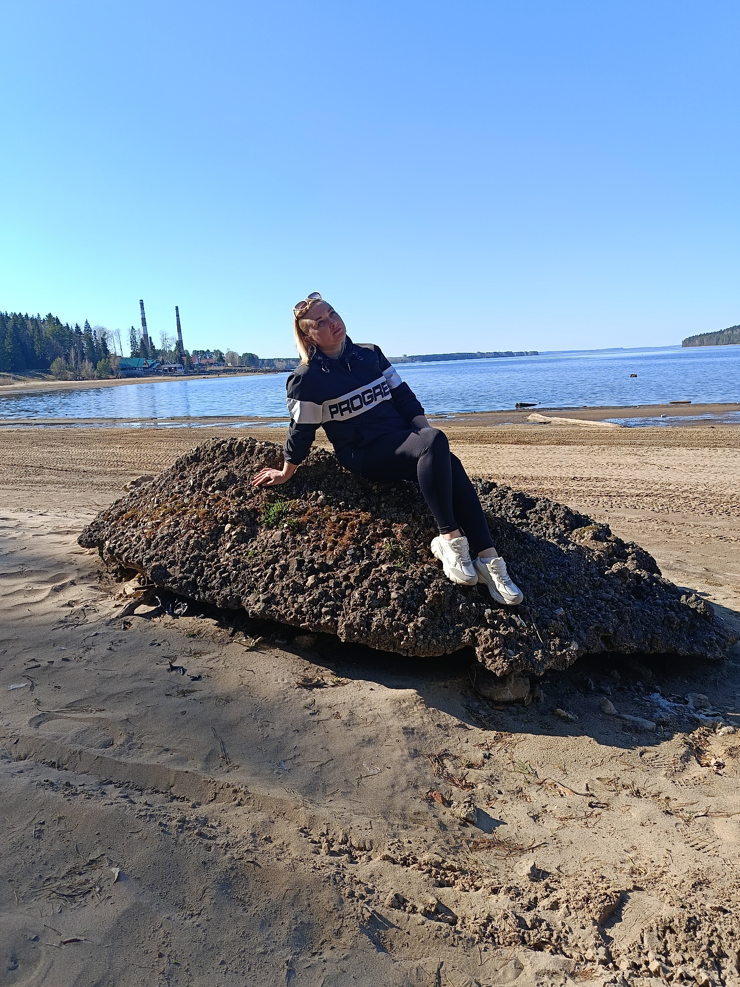 A woman sits on a large rock on the sandy beach in Chaikovsky, Russia. The city is visible in the background, with industrial buildings and smokestacks. The water is calm. The woman is wearing a dark jacket, black leggings, and white sneakers. She appears relaxed and is leaning on the rock. The sky is clear and blue. The beach is sandy with some dry vegetation.